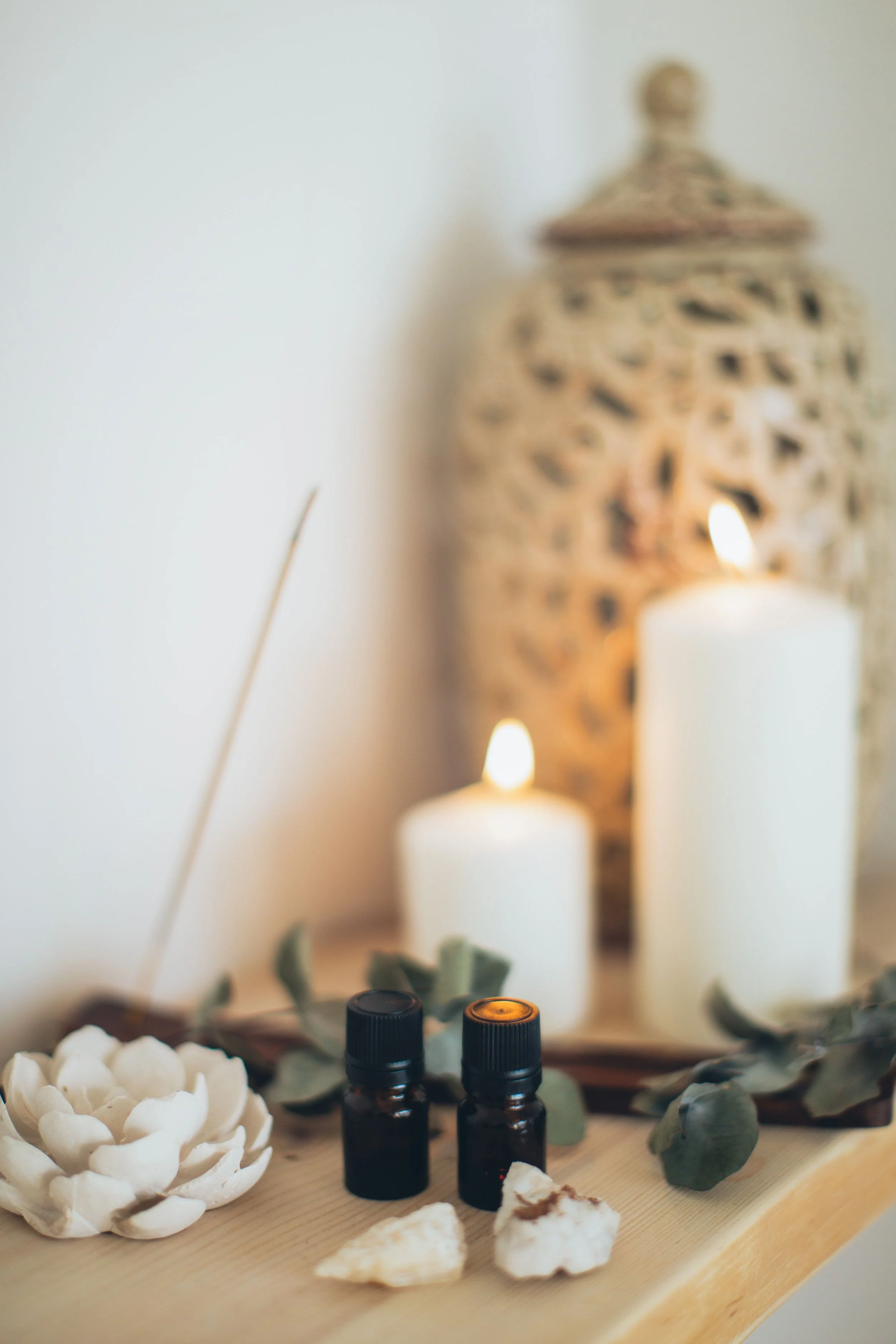 Velas aromáticas, aceites esenciales y piedras decorativas sobre una bandeja de madera, con fondo difuso de un adorno de cerámica y velas encendidas.