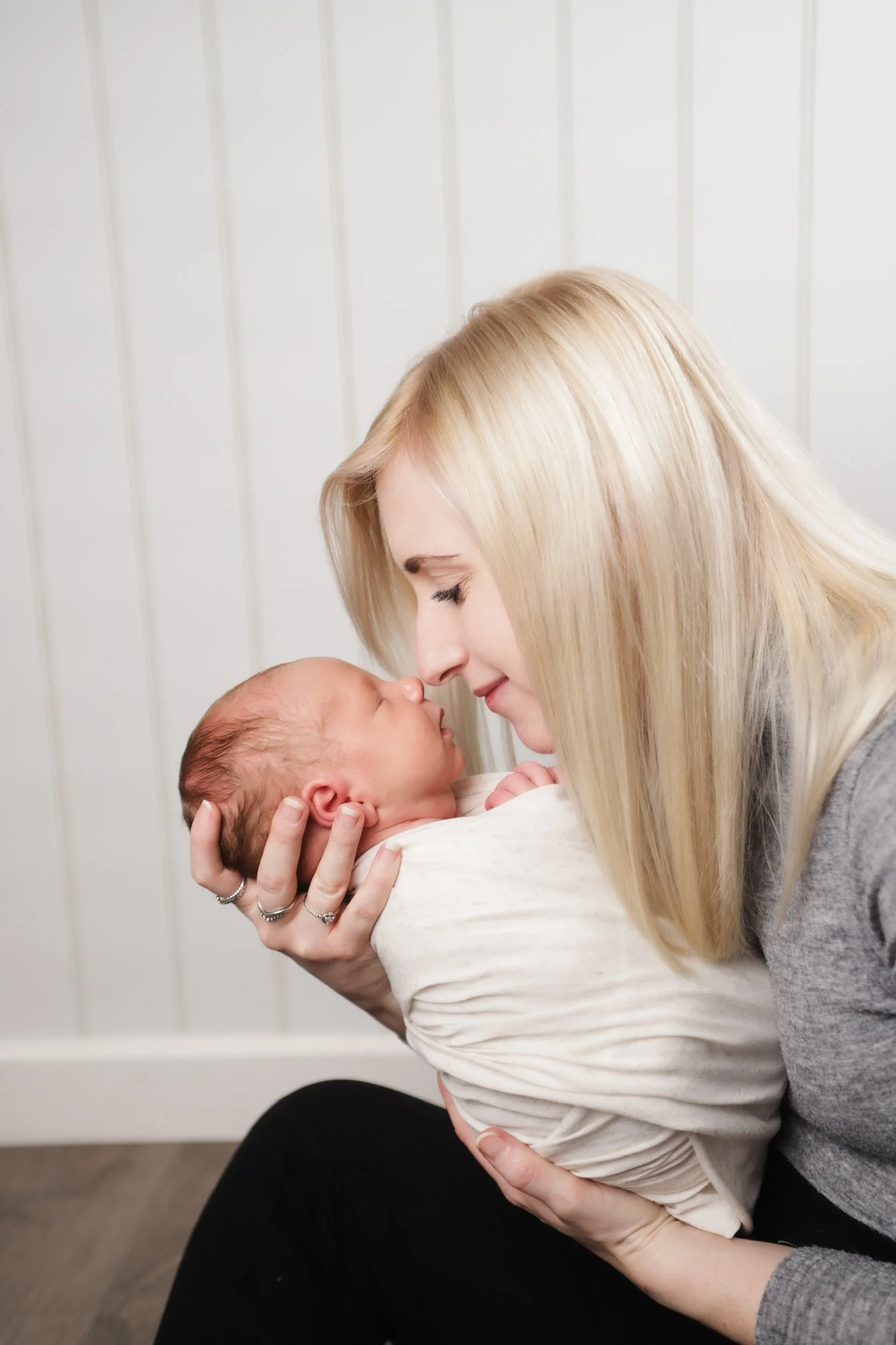 A woman with long blonde hair gently holding a newborn baby, with both facing each other affectionately. The woman is sitting against a white paneled wall.