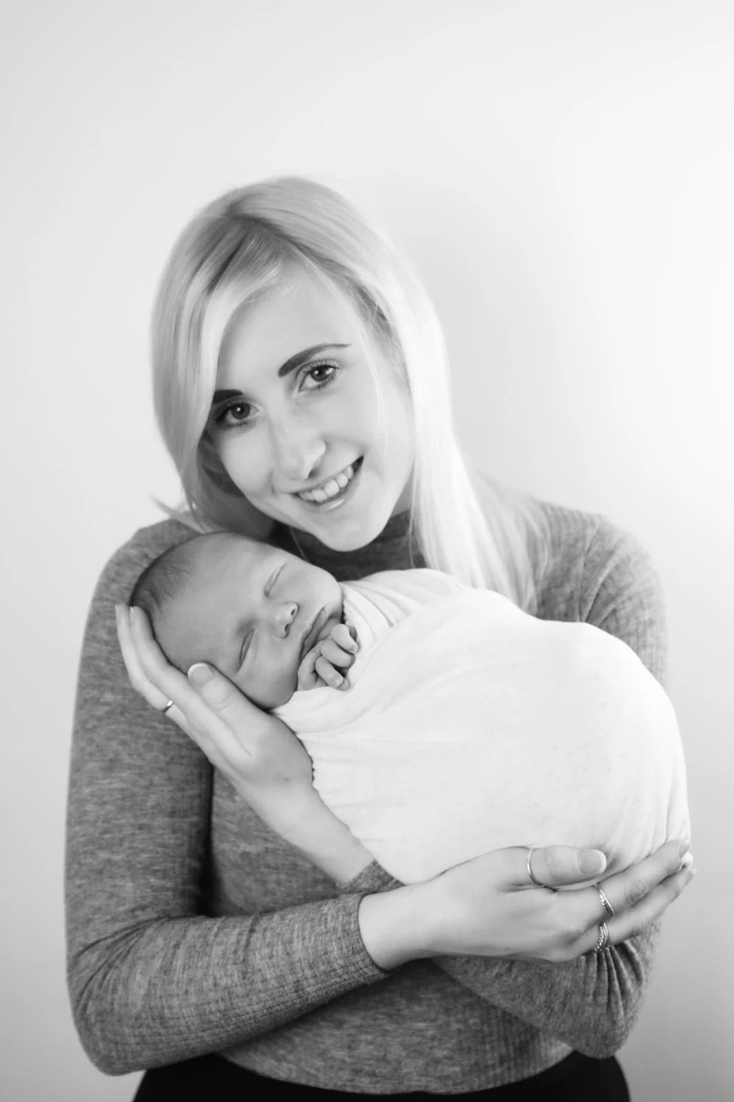 A woman with long blonde hair smiling and holding a sleeping newborn baby wrapped in a blanket.