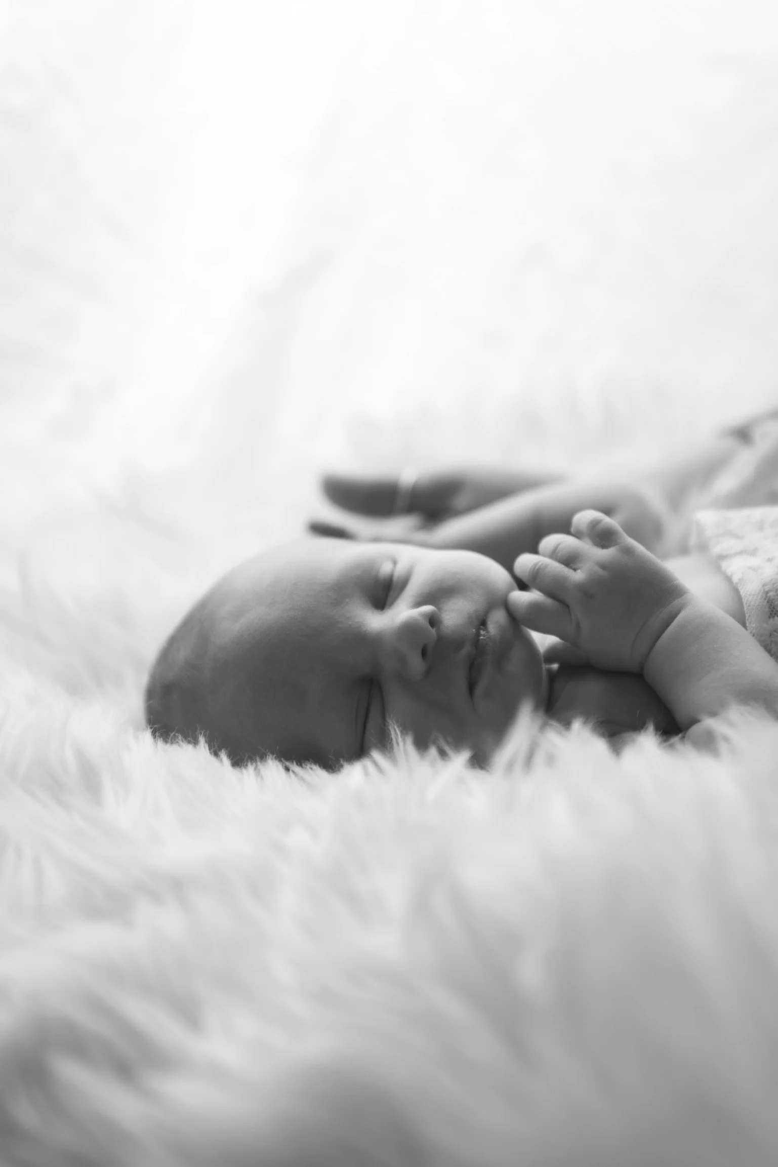 A newborn baby lying on a soft, fluffy surface, with eyes closed and hand near the face, in black and white.