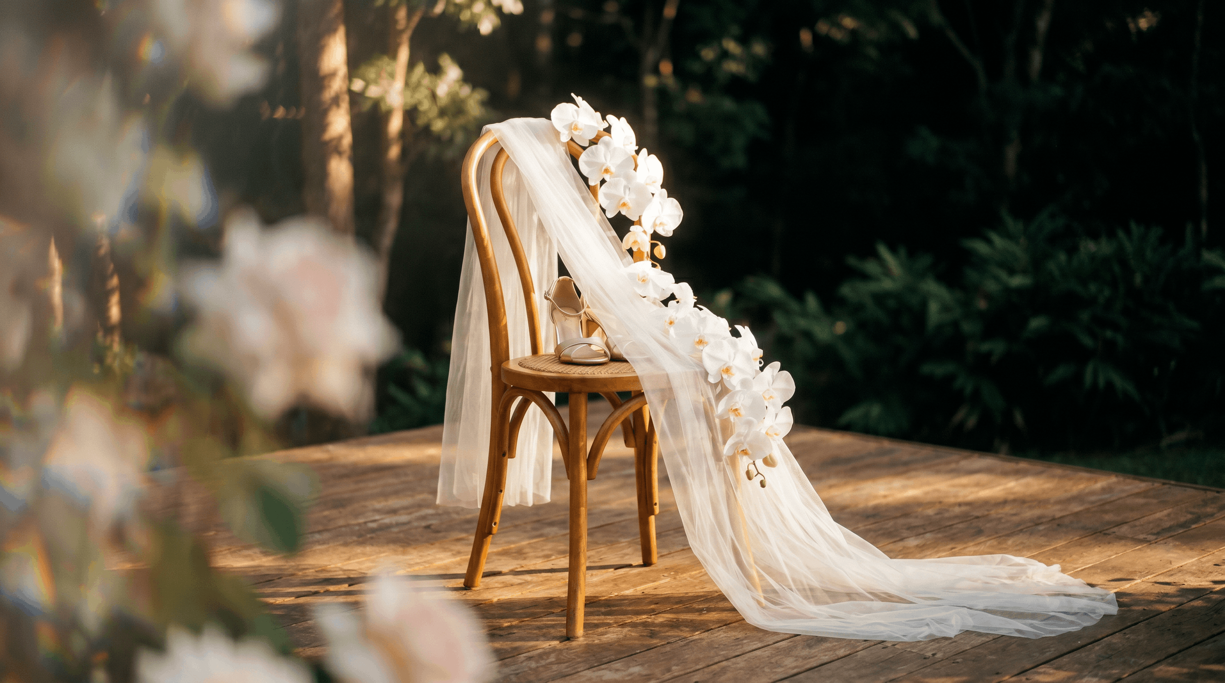 A wooden chair decorated with white flowers, a sheer white fabric, and a pair of beige high heels, set on a wooden platform outdoors with greenery in the background.