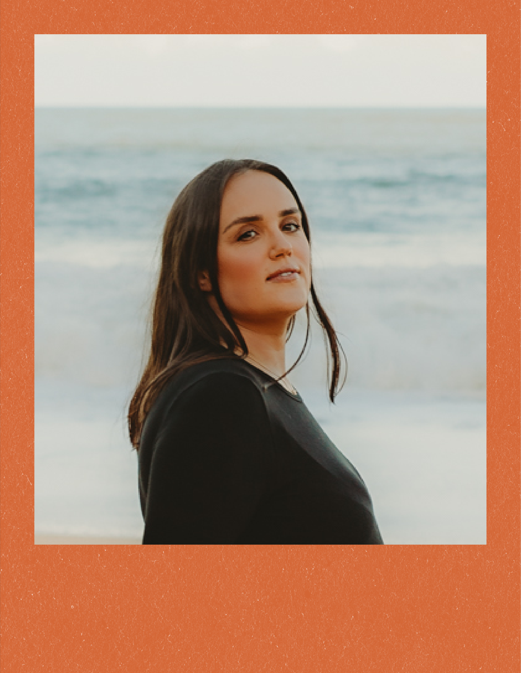 A woman with long dark hair and light skin is standing outdoors near the ocean with the sky above. She is wearing a black top and is looking towards the camera with a slight smile.