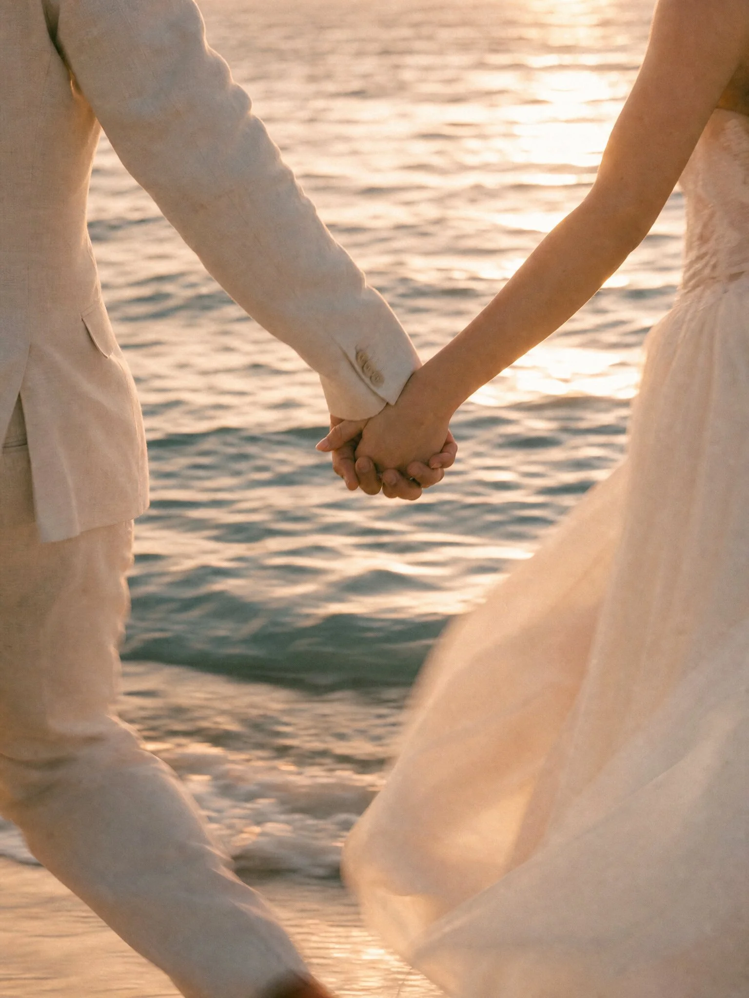 A couple holding hands at the beach during sunset, with their lower bodies and intertwined hands visible, surrounded by sand and water.