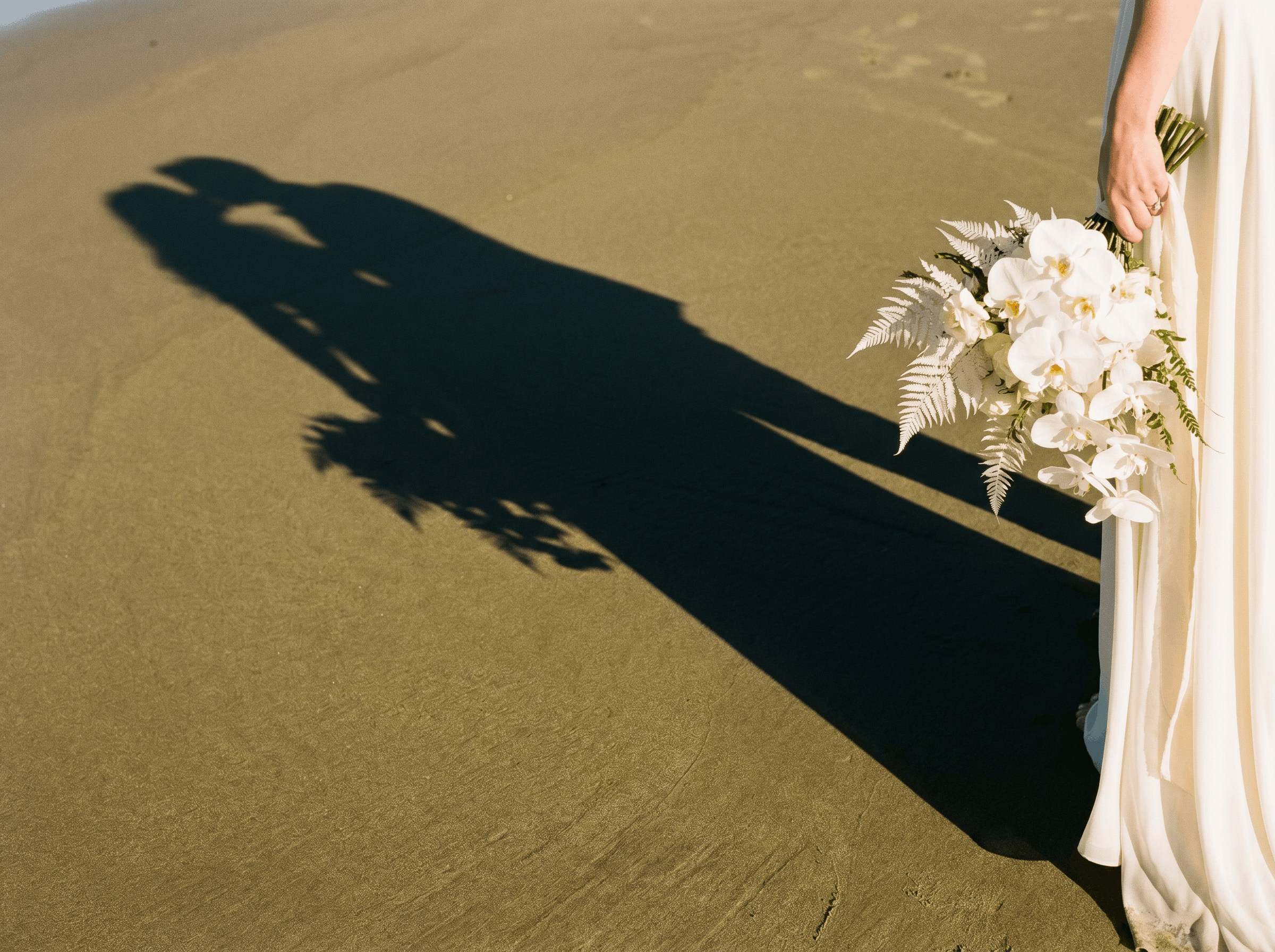 A wedding dress with a bouquet of white orchids and ferns held by a person standing on a concrete floor, casting a shadow.
