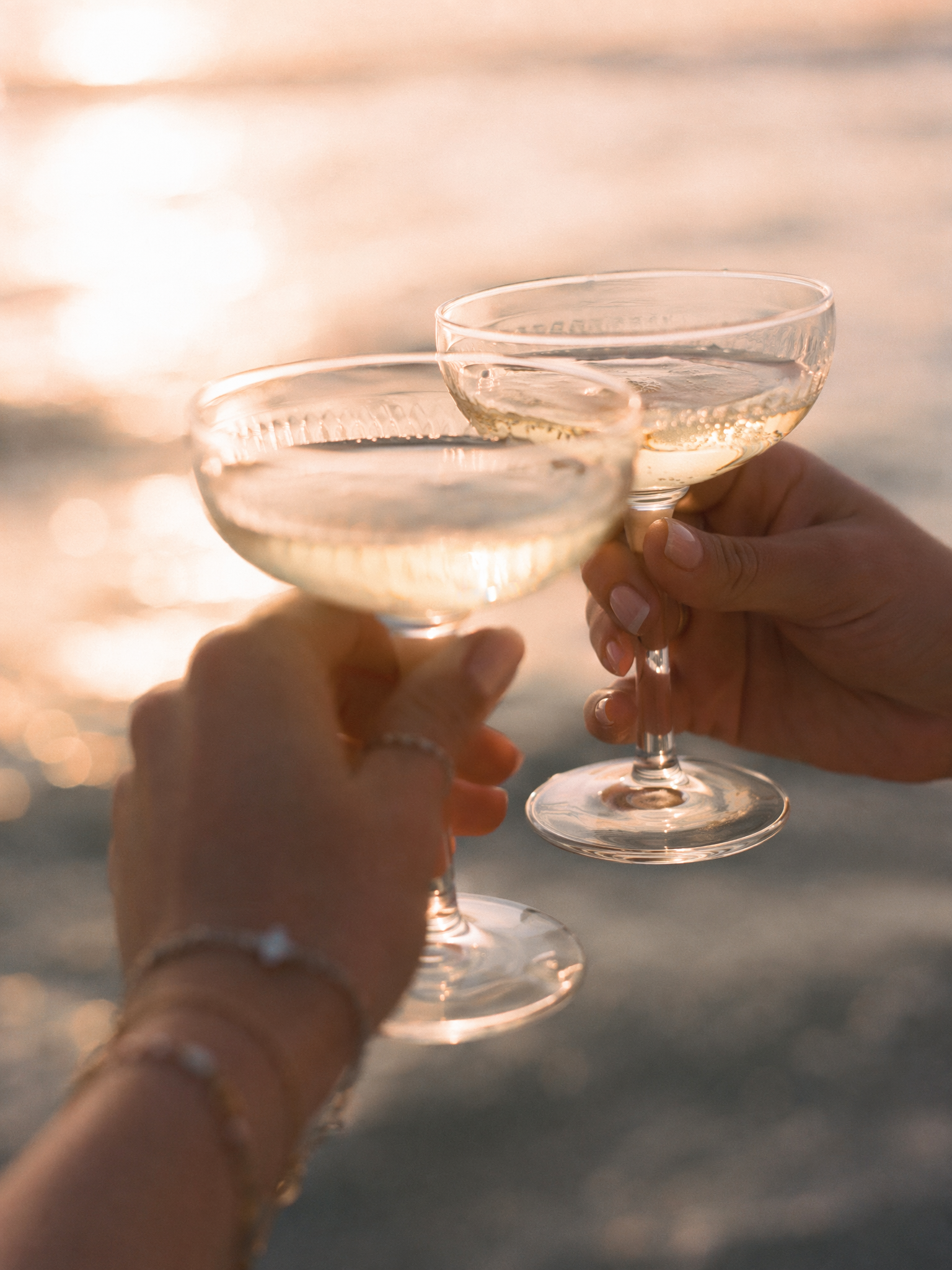 Two hands holding champagne glasses clinking together on a beach at sunset.