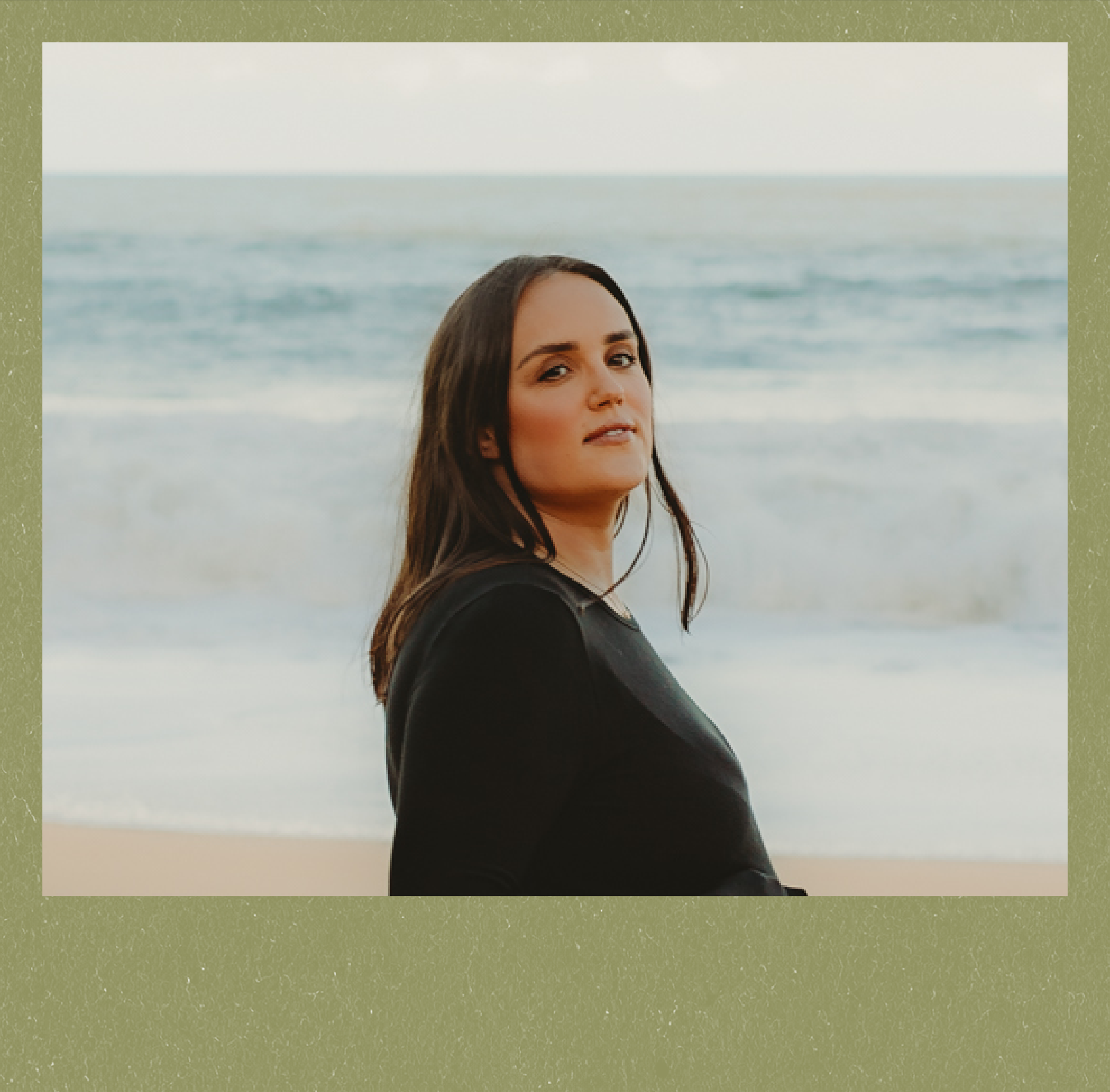 A young woman with long dark hair standing on a beach near the ocean, wearing a black top, looking slightly over her shoulder with a calm expression.