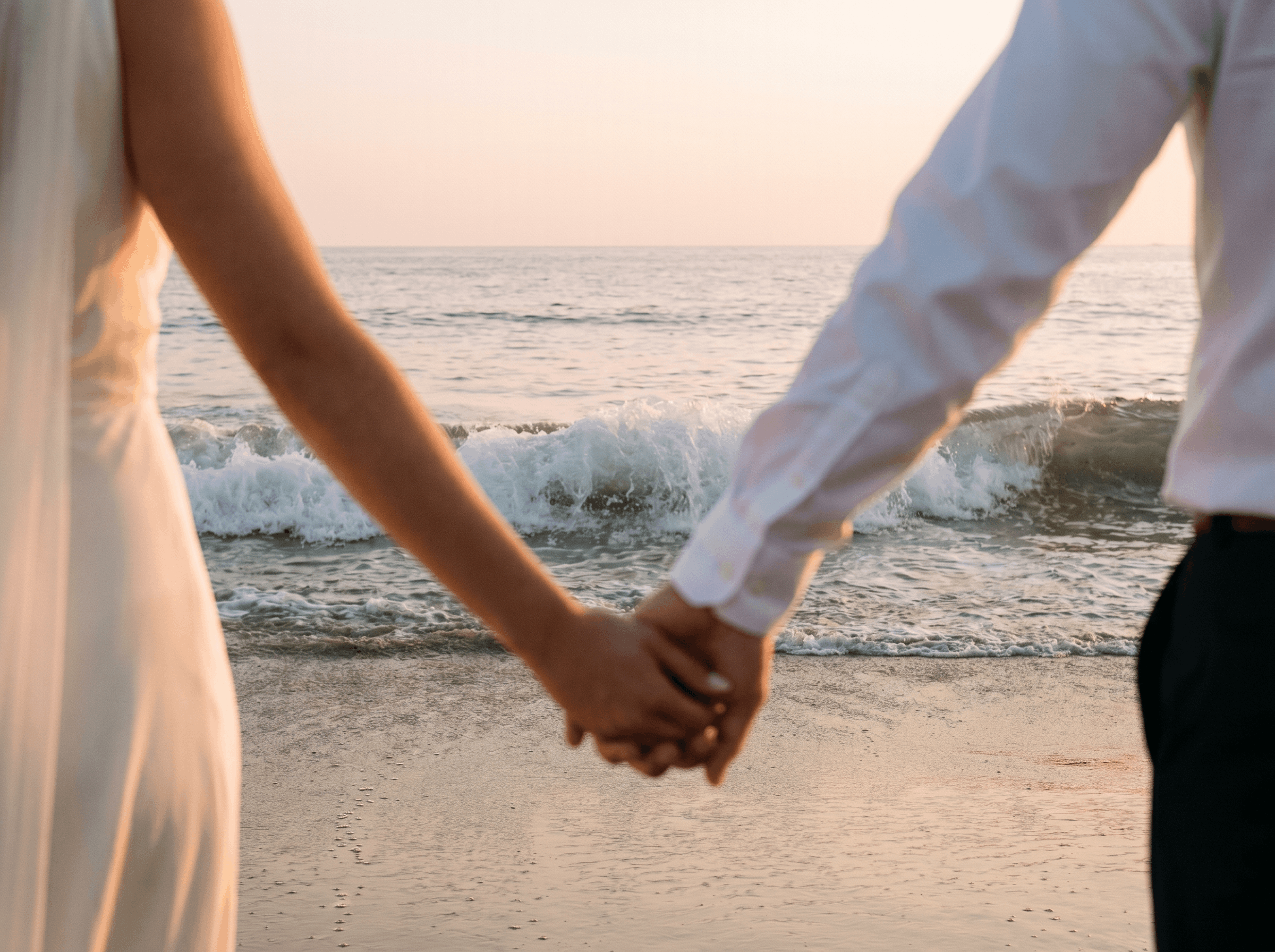 A close-up of a couple holding hands on the beach at sunset, with the ocean and sky in the background.