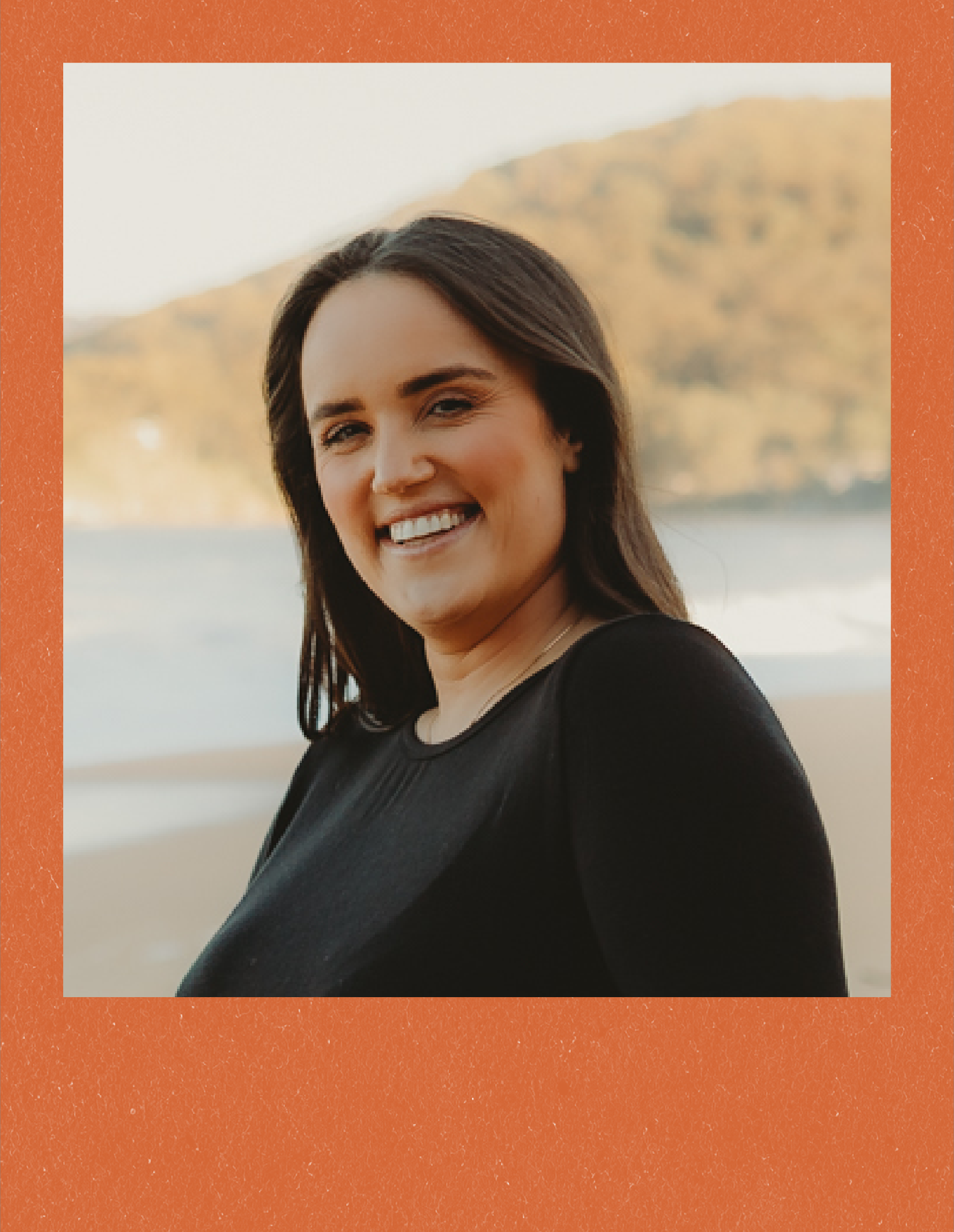 A young woman with dark hair smiling outdoors near a water body, with a mountain and clear sky in the background.