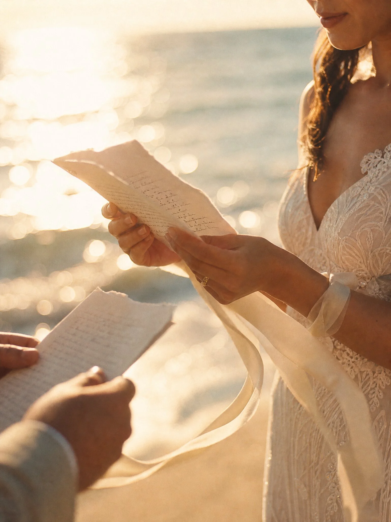 A woman in a white lace wedding dress reading a handwritten letter during a sunset by the water with sunlight reflecting off the surface.