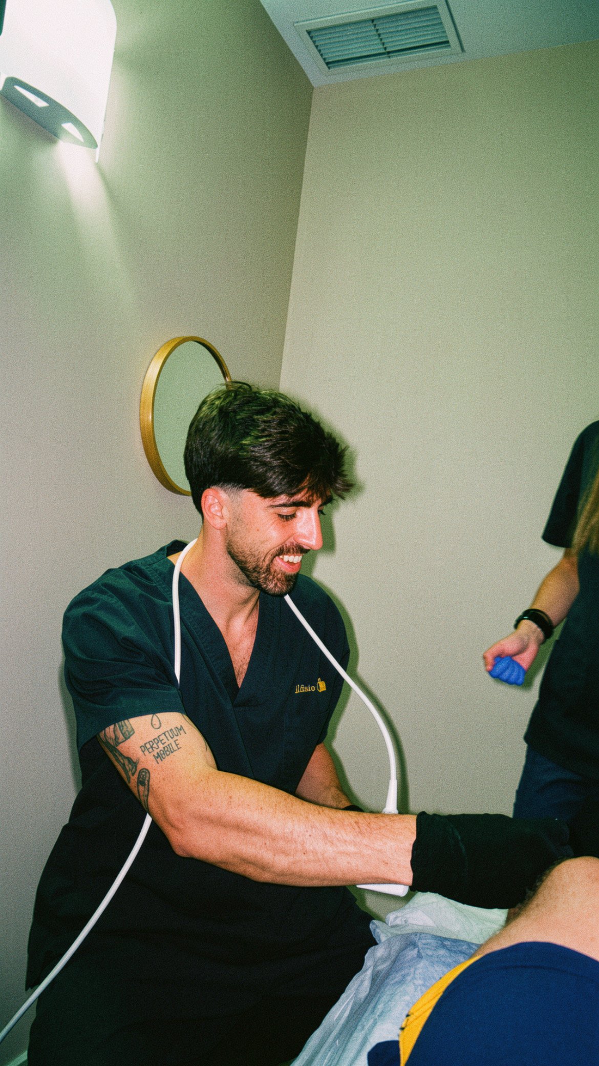 Un médico sonriente revisa a un paciente en un consultorio, con un estetoscopio alrededor del cuello y tatuajes en el brazo.