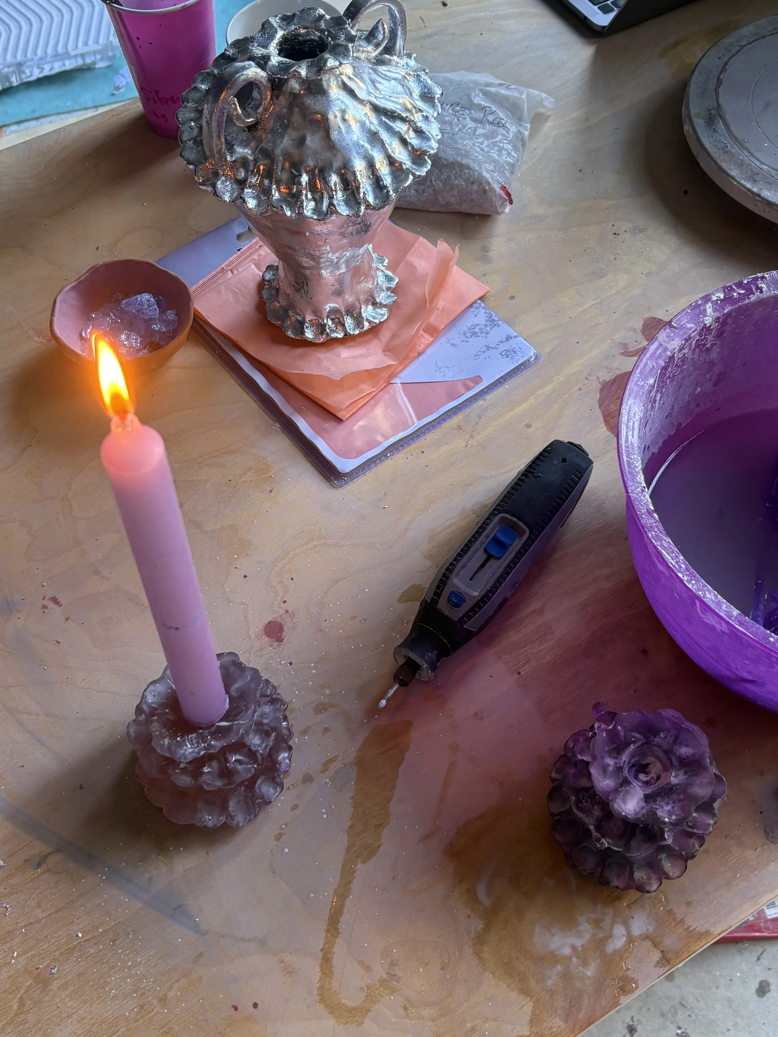 A workspace with a silver decorative vase, a pink candle, a pink cup, a plastic bag, a purple bucket with water, a remote control, and a small power drill, with spilled water on the wooden table.