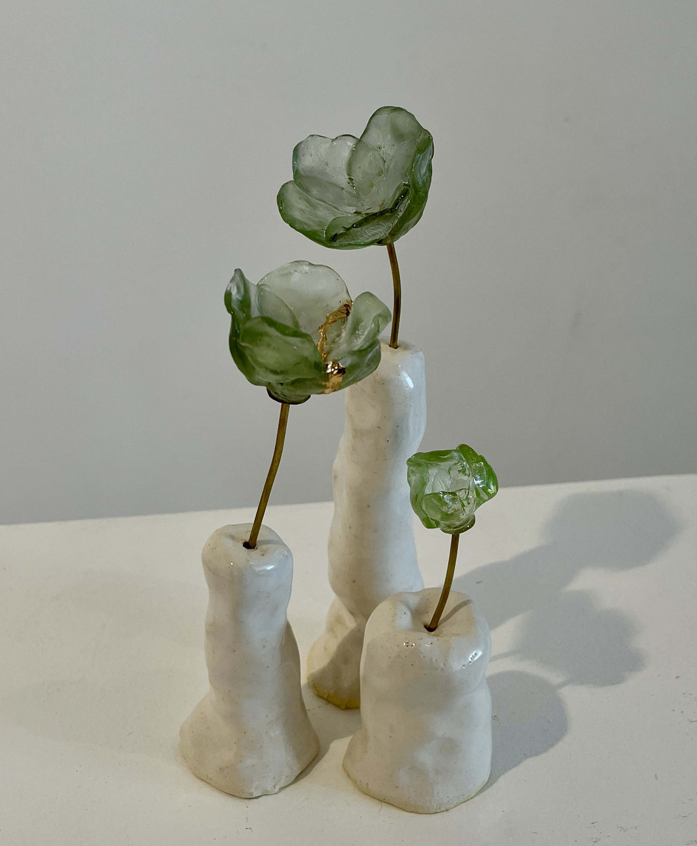 A collection of small white sculptures resembling abstract human figures with elongated shapes, topped with green glass flowers. The sculptures are arranged on a white surface against a plain white background.