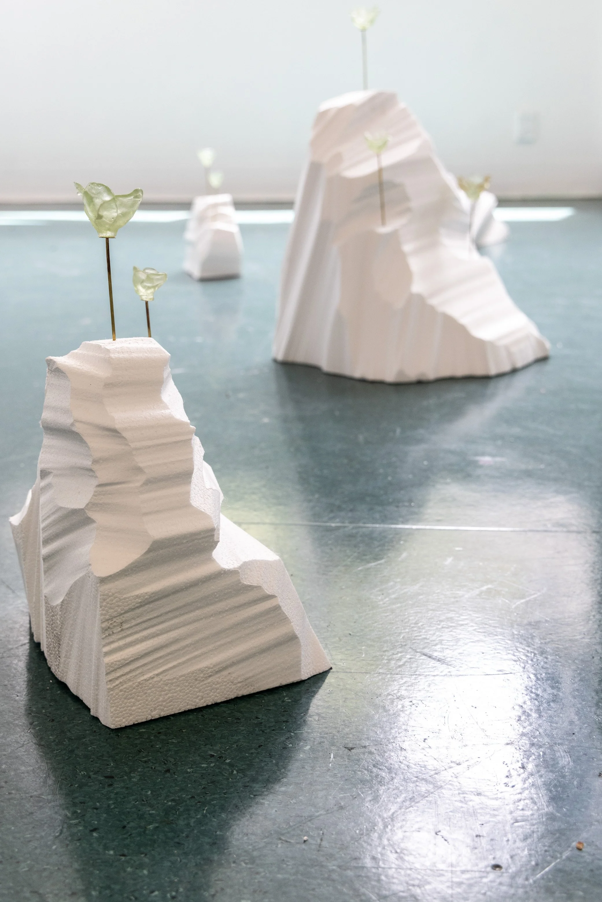 White rock formations with carved layers, some topped with green glass flowers on thin metal sticks, placed on a greenish floor