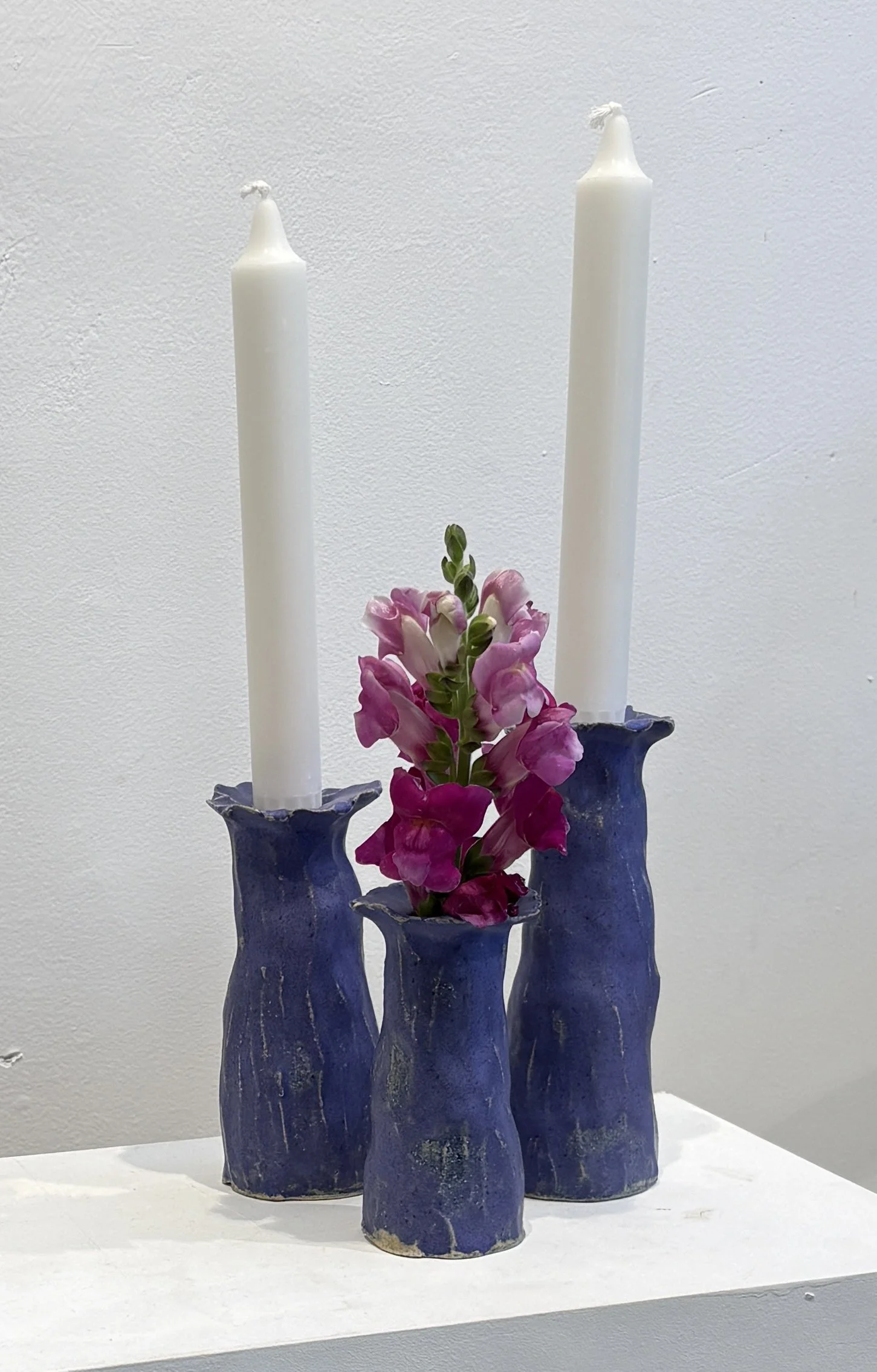 A decorative arrangement of three dark blue ceramic vases holding pink flowers and two tall white candles against a plain white wall.