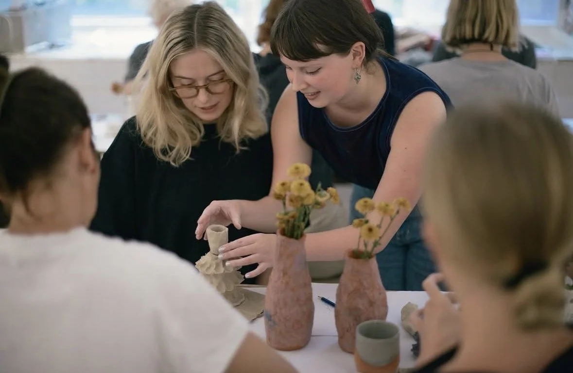 Two women demonstrating pottery to a group of women seated at a table in a well-lit room.