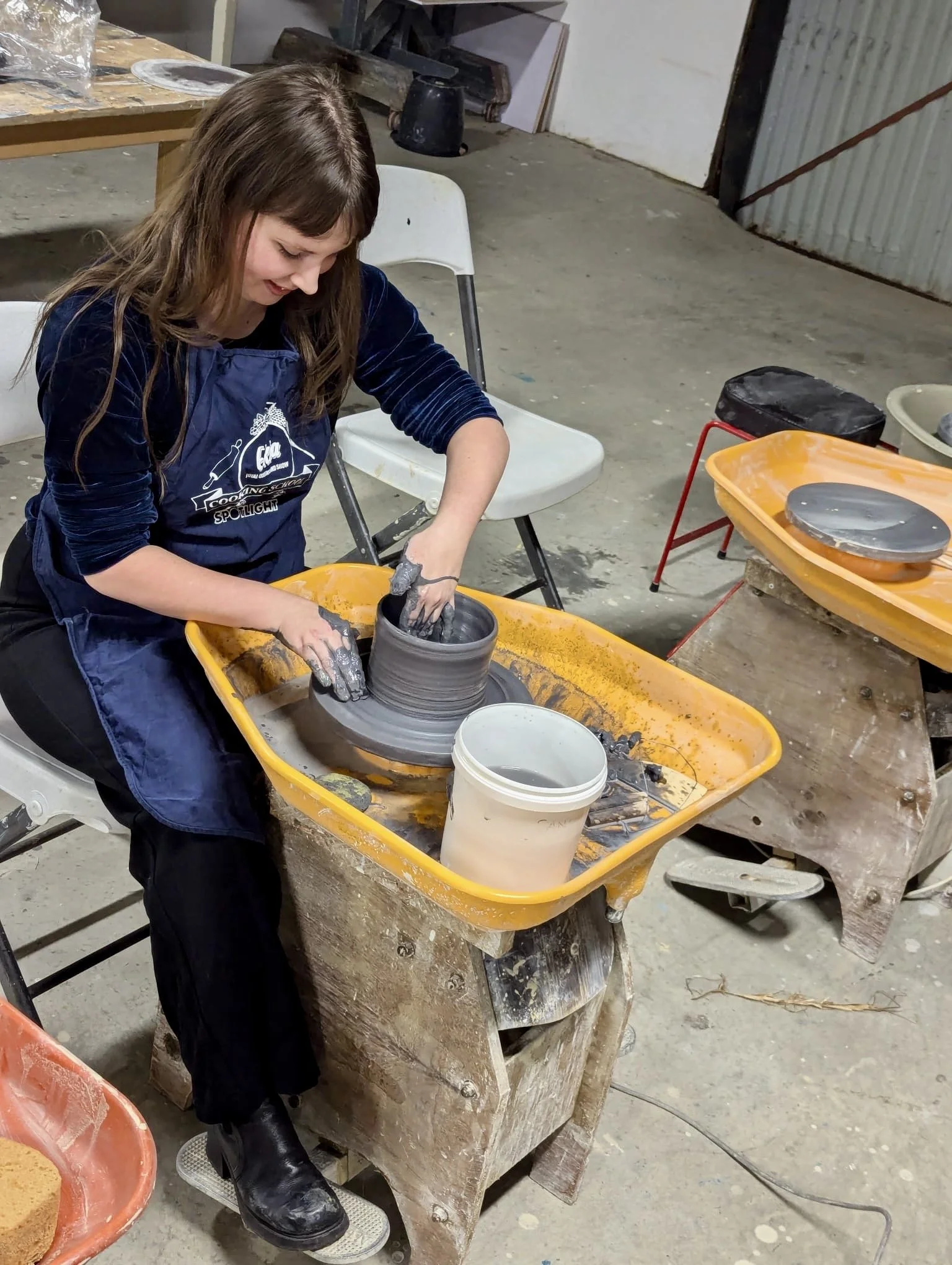 A woman creates pottery on a pottery wheel in a workshop. She is shaping gray clay with her hands, wearing a black apron, and has long brown hair. There are various tools and containers of clay around her on the table and wheel.