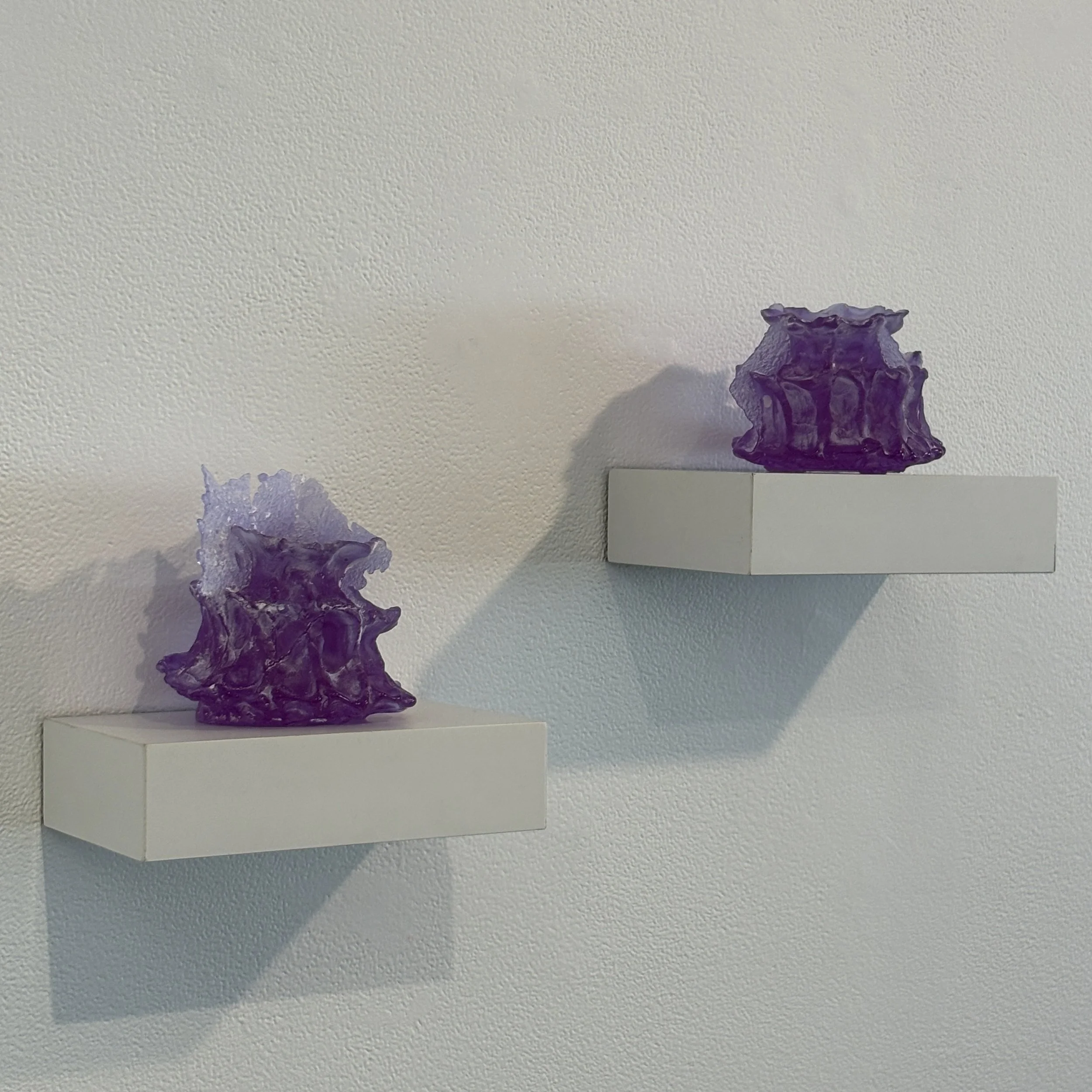 A textured white wall with two purple glass sculptures placed on white platforms. The sculptures are abstract and irregular in shape.