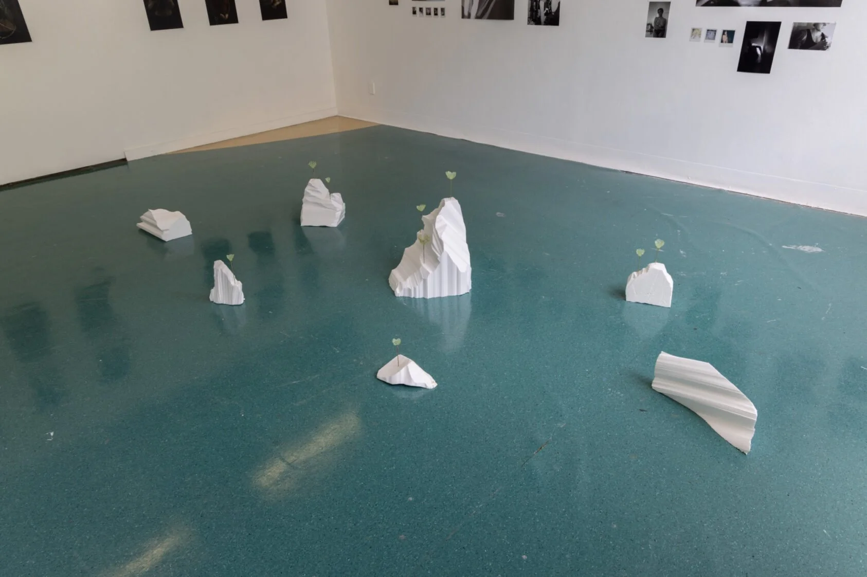 Indoor art installation featuring several white paper sculptures resembling mountains on a green floor, with small green paper hearts attached to some of the peaks, and black and white photographs on the wall in the background.