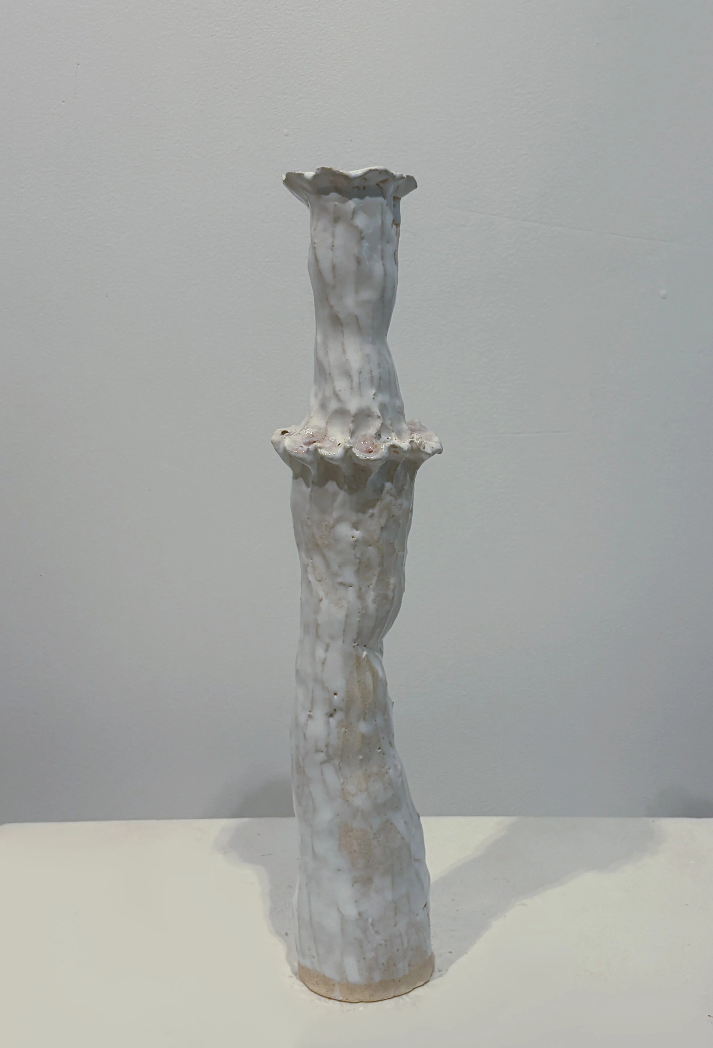 A tall, white ceramic vase with a textured, uneven surface and wide, scalloped rim, standing on a white surface against a plain, light-colored background.