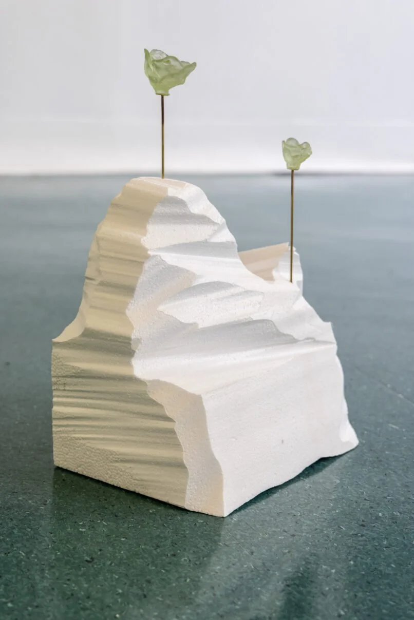 White abstract sculpture resembling a mountain or iceberg with two thin metal rods topped with green translucent leaf-like shapes.