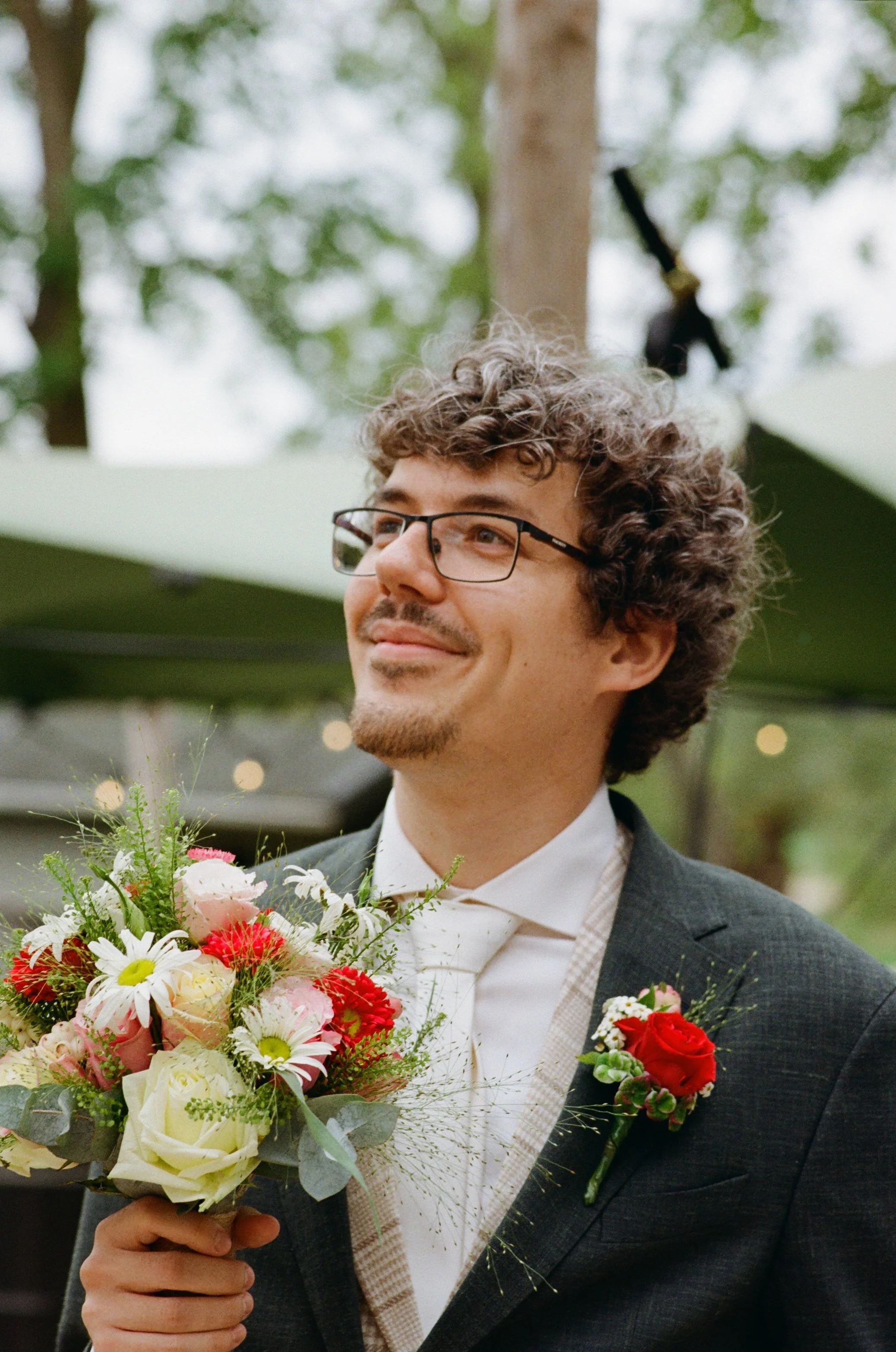 A man with curly hair, glasses, and a beard wearing a wedding suit and holding a bouquet of flowers at a wedding celebration.