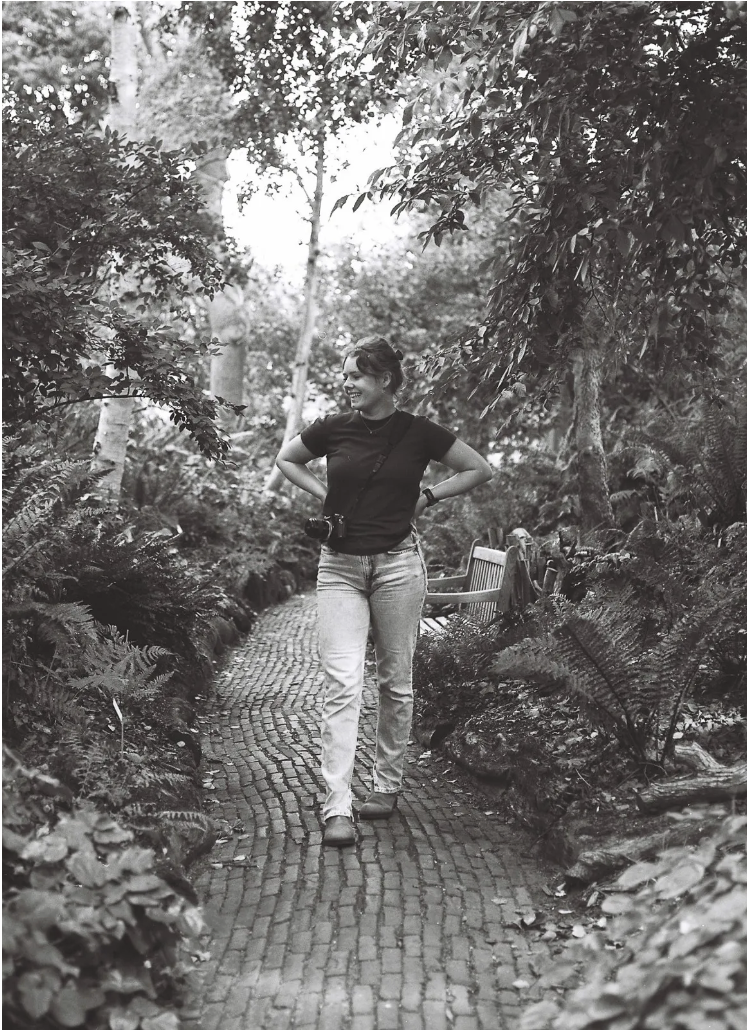 A woman walking along a cobblestone path in a lush, wooded garden, smiling and carrying a camera around her neck.