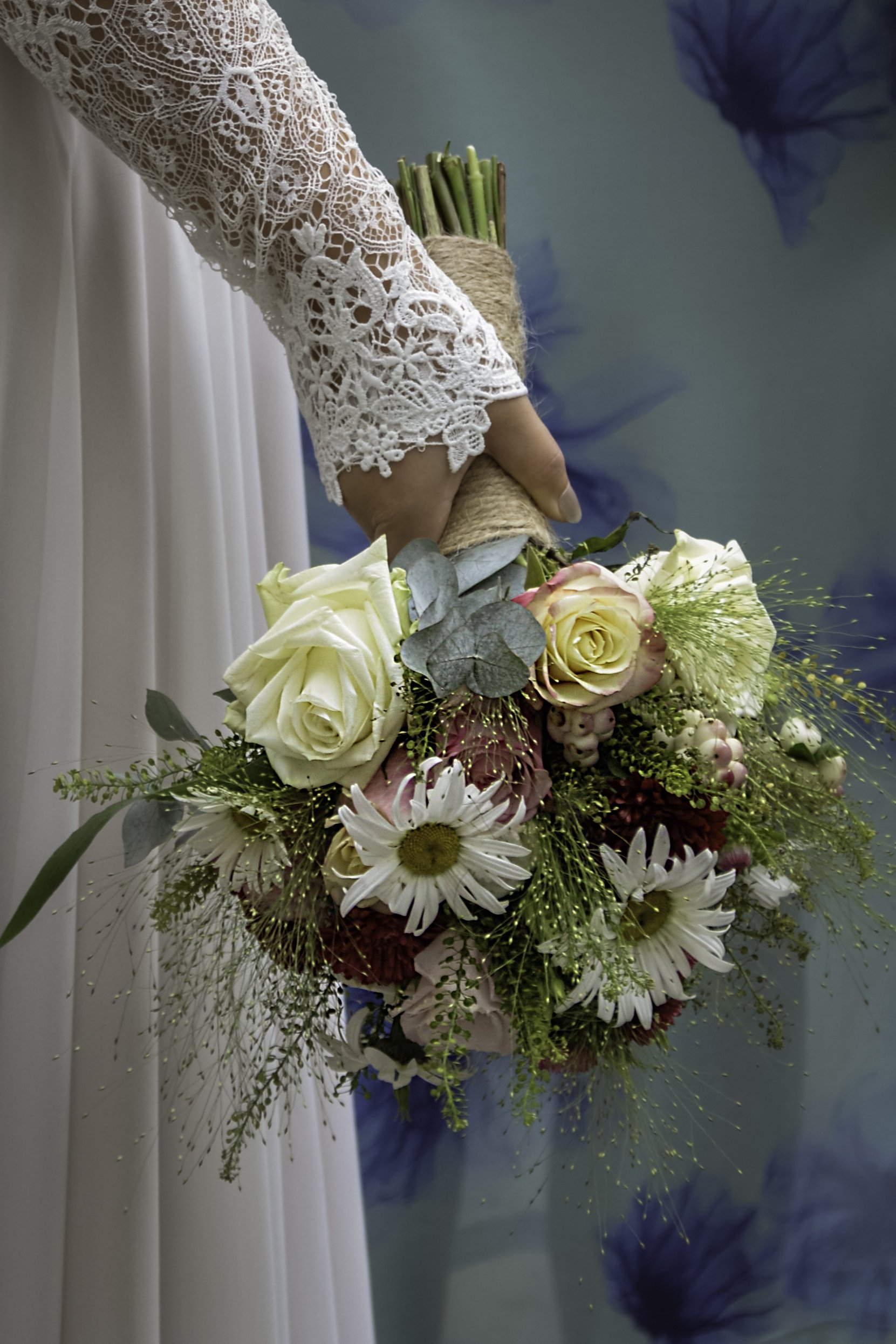 Close-up of a person wearing a white lace dress holding a large bouquet of flowers, including roses, daisies, and greenery, against a backdrop of blue and white fabric.