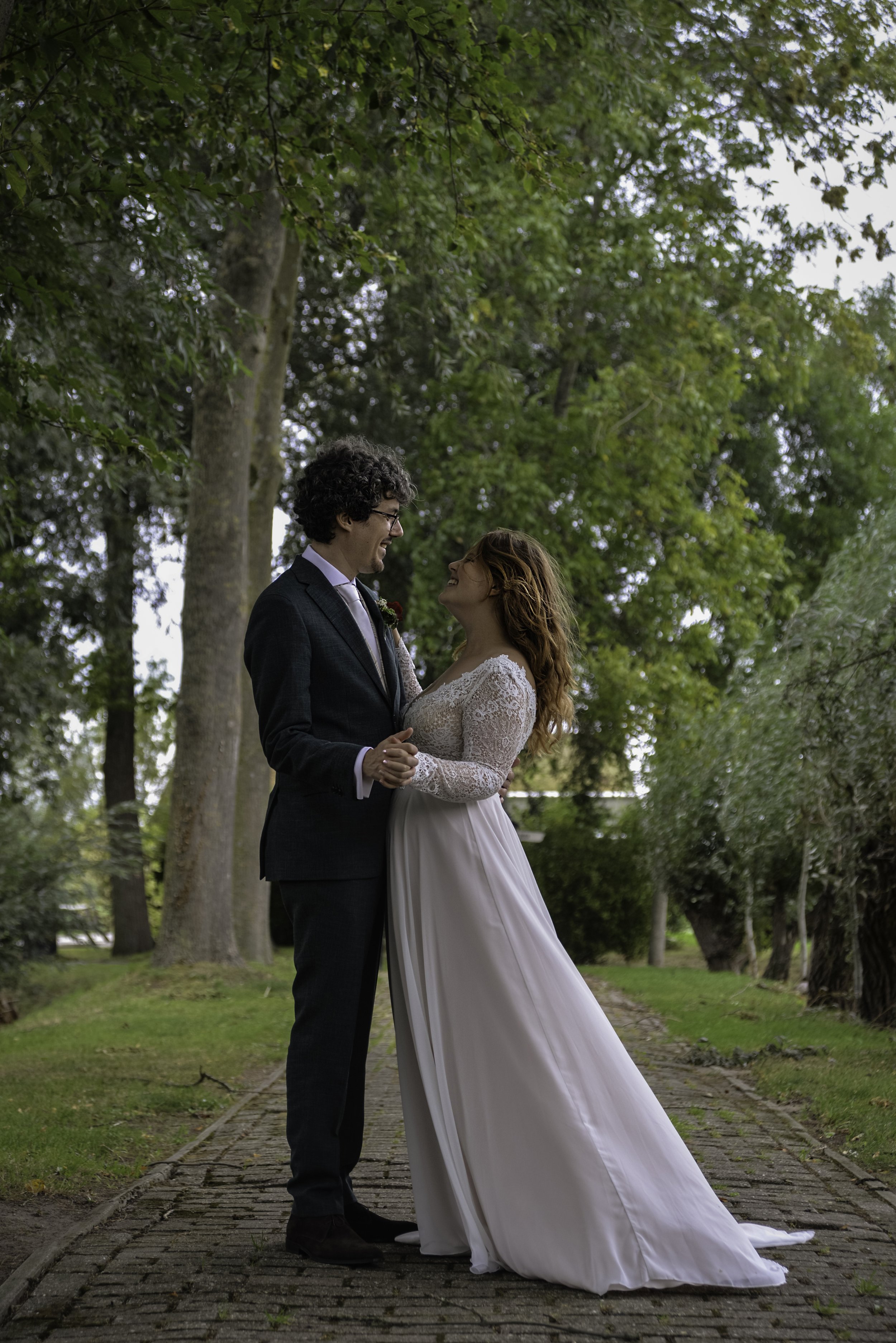 A bride and groom standing close, holding hands, smiling, in a park with tall trees, a brick path, and green grass, during daytime.