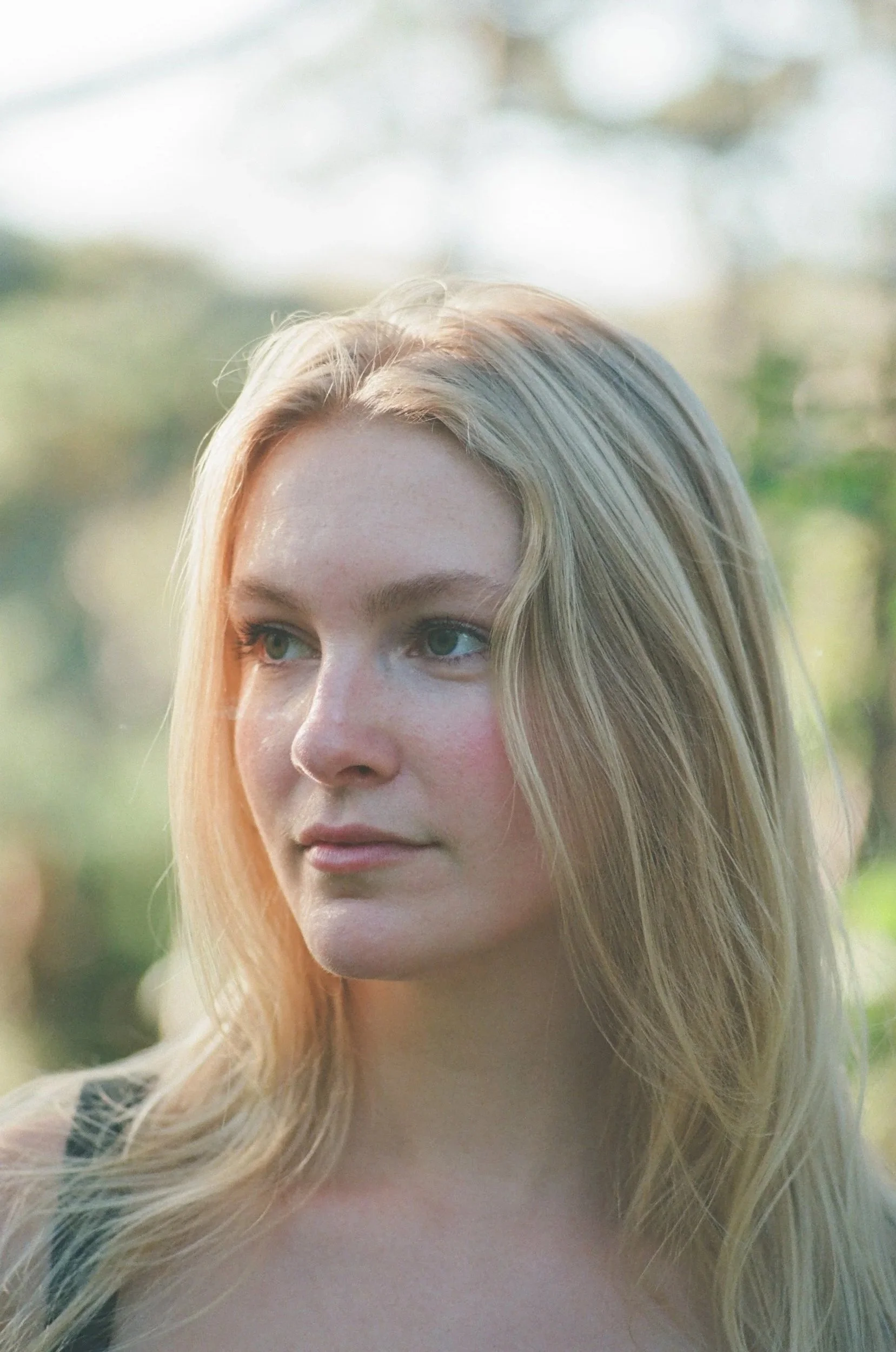 Close-up of a woman with long, blonde hair outdoors with a blurred background of trees and sky.