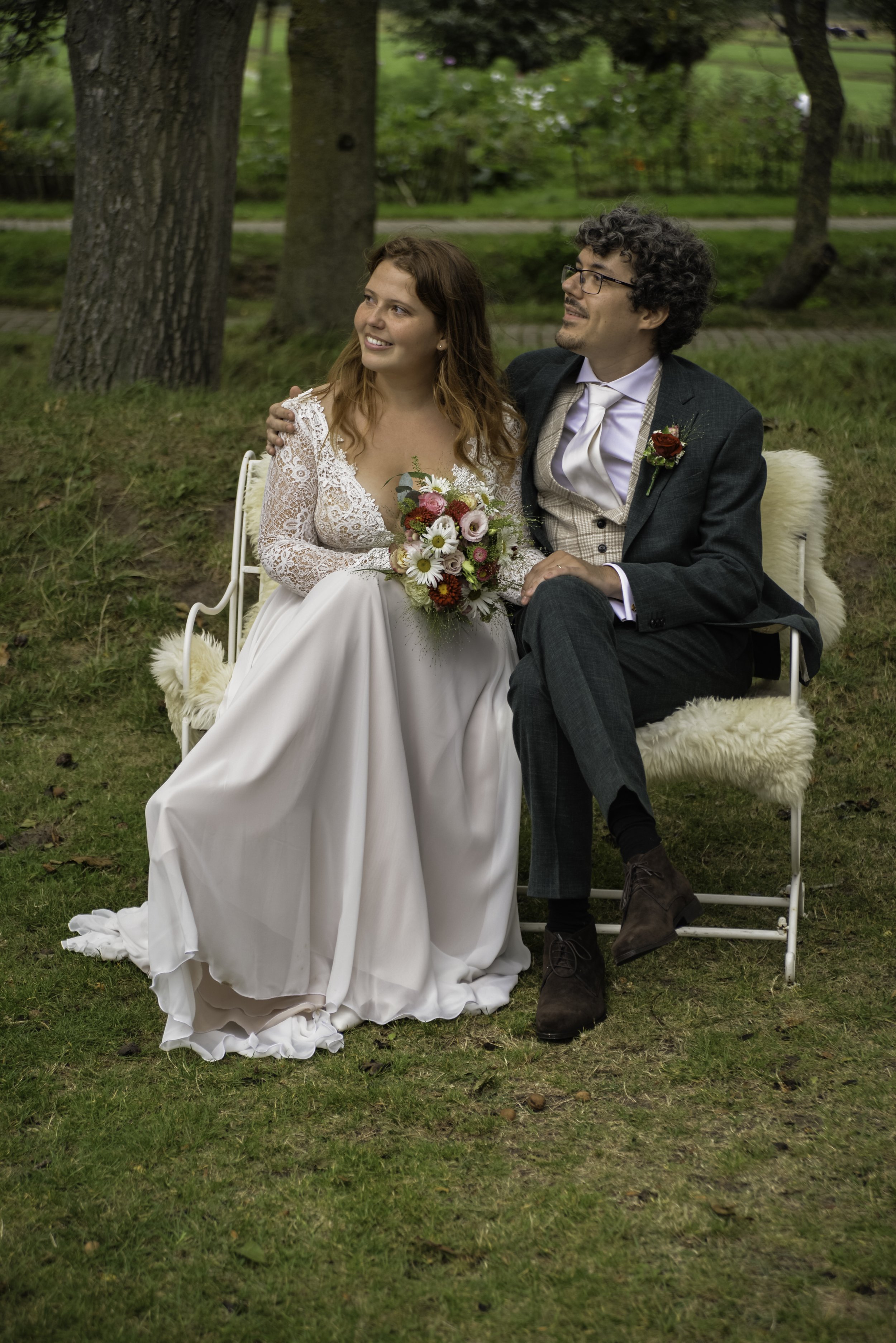 A newlywed couple sitting together outdoors on a bench in a park with trees and grass in the background. The bride wears a white lace wedding dress with long sleeves and holds a colorful bouquet. The groom is dressed in a dark green suit with a tan v