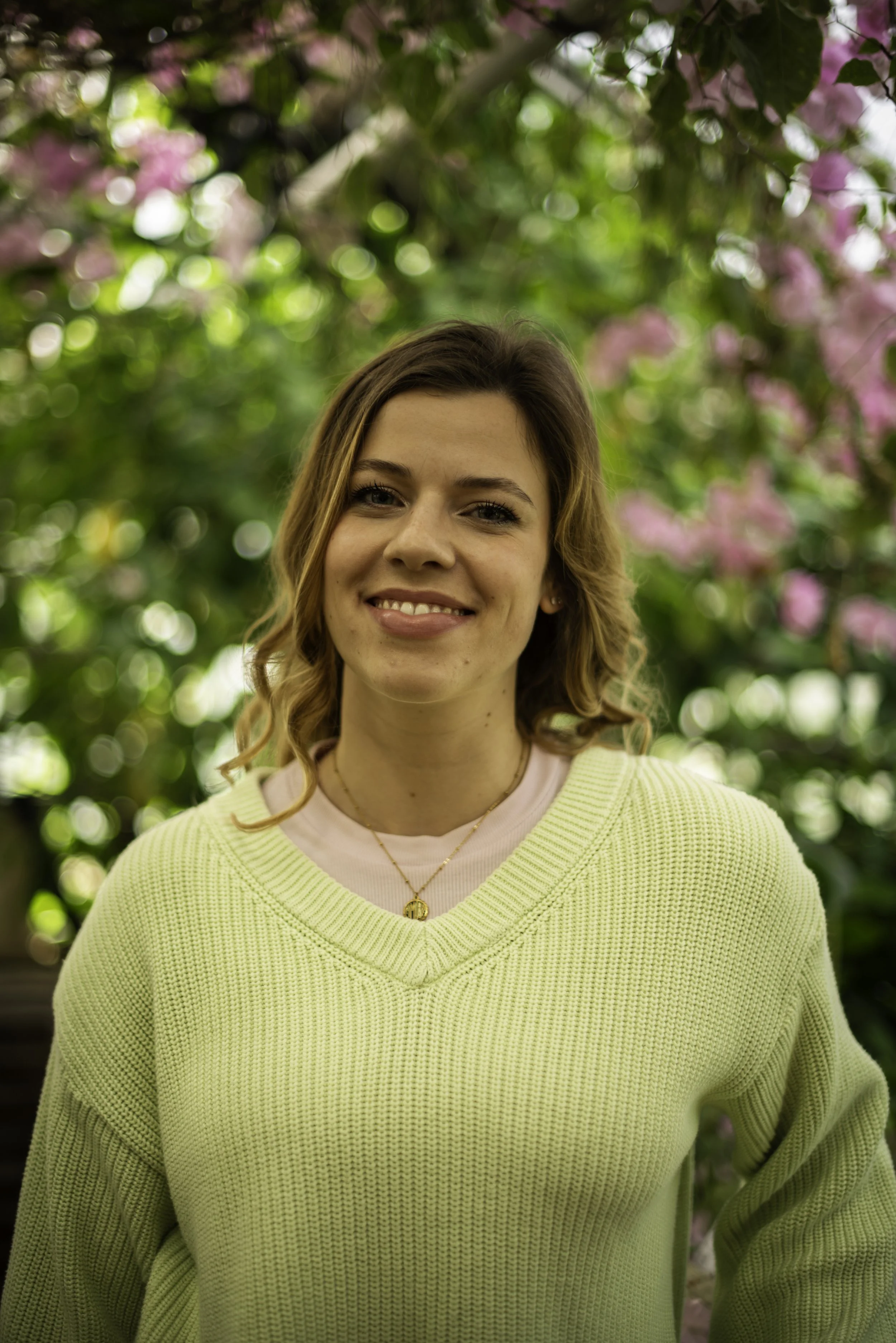 A woman with light brown hair styled in loose waves, smiling, wearing a light pink shirt underneath a yellow knitted sweater, standing outdoors with pink flowers and green foliage in the background.