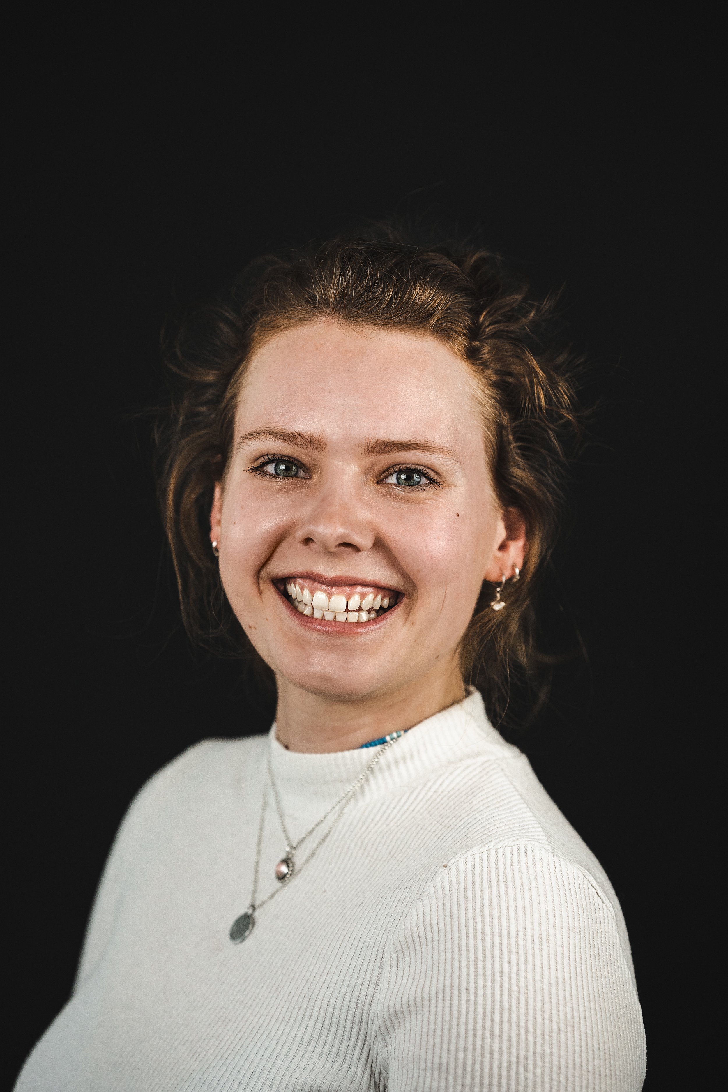 A smiling woman with curly blonde hair, green eyes, and fair skin, wearing a white ribbed turtleneck sweater, layered necklaces, and earrings, against a plain black background.