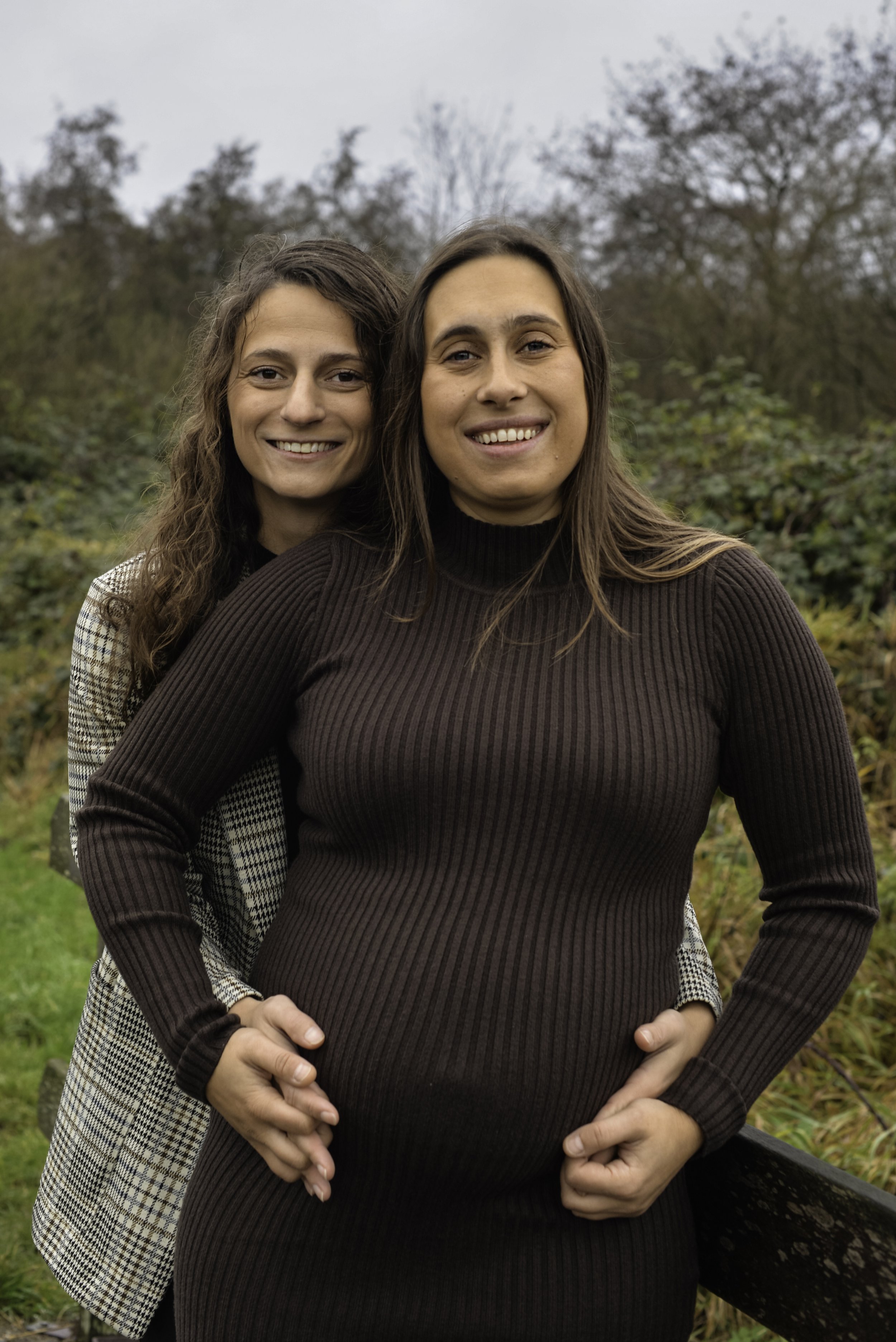 Two women standing outdoors in a park, one in front with hands on hips and the other behind with arms around her shoulders, both smiling and looking at the camera.