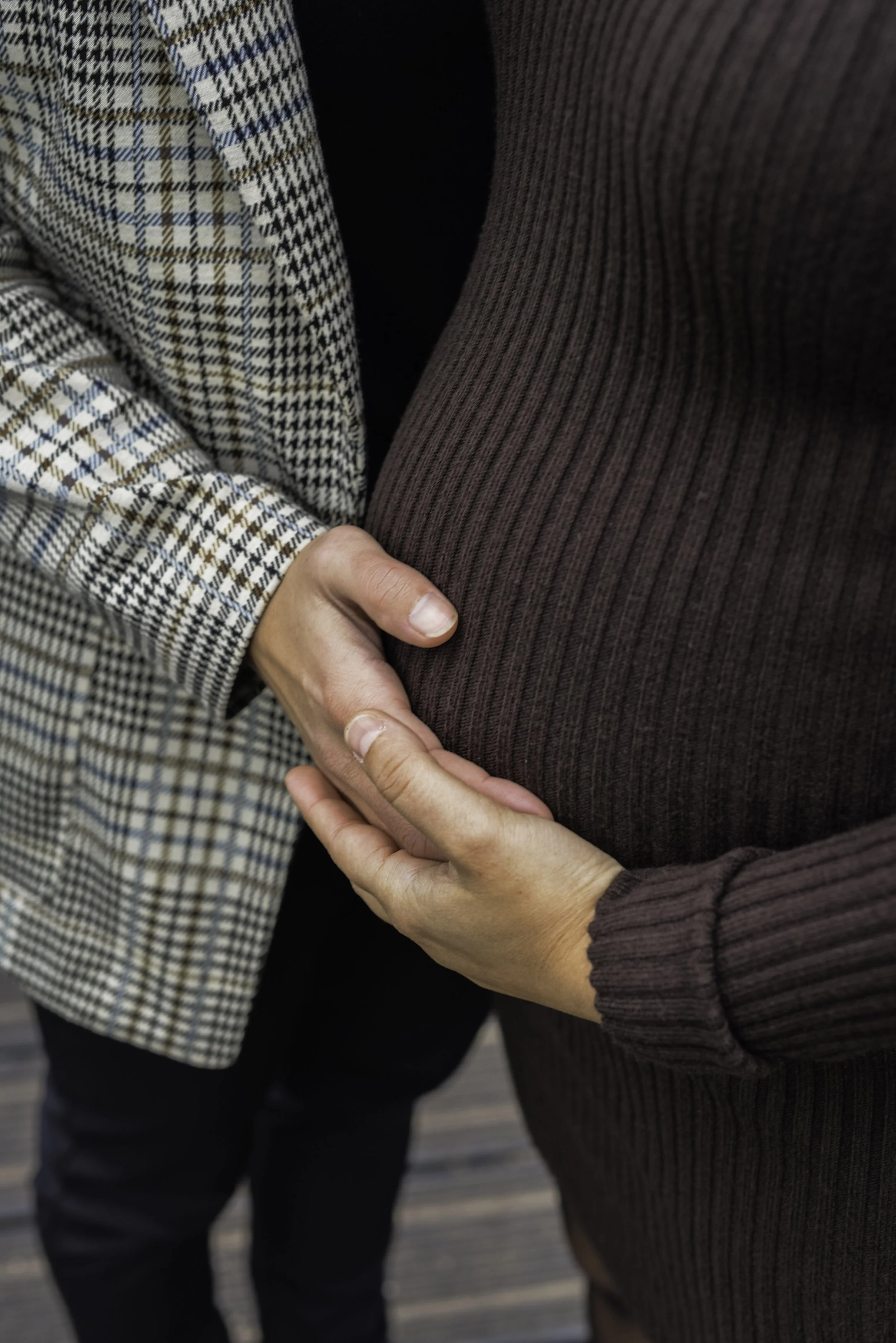 A person is gently placing their hand on another person's pregnant belly. Both individuals are wearing long-sleeved, patterned clothing; one in a checked blazer and the other in a ribbed knit top.