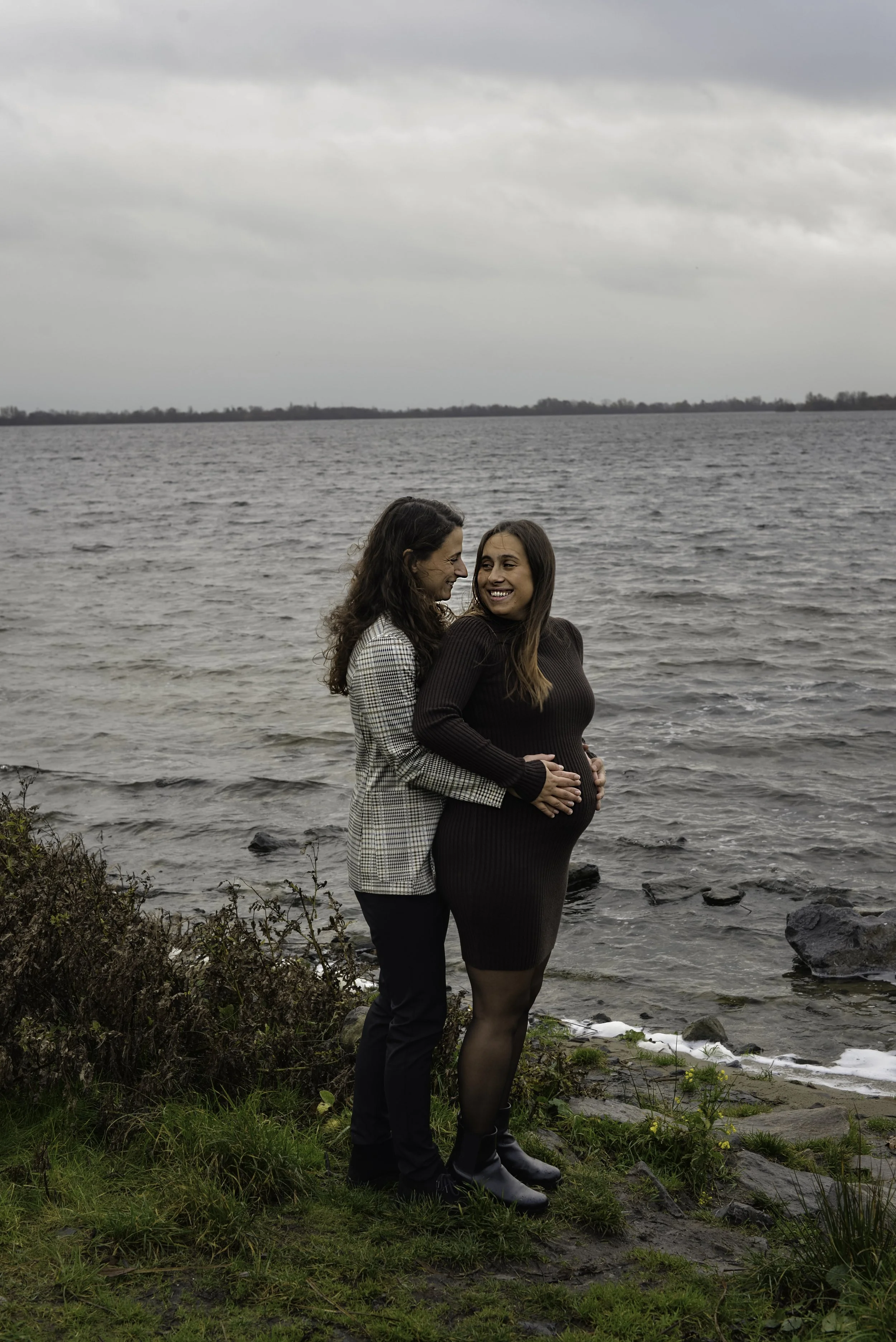 A woman in a black dress and high heels with pregnant belly, standing close to her partner at the lakeside, both smiling and looking at each other. The man is dressed in dark pants and a checkered blazer, and they are embracing among rocks and grass 