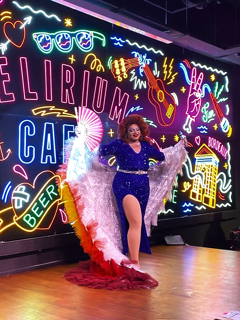 Une personne déguisée avec une robe bleue brillante et une cape colorée se tient sur scène devant une affiche de néons représentant la ville de Bordeaux, avec des éléments comme une guitare, un verre de bière, un soleil, et des lettres comme 'FELIRIUM', 'CAPE', 'BEER'.
