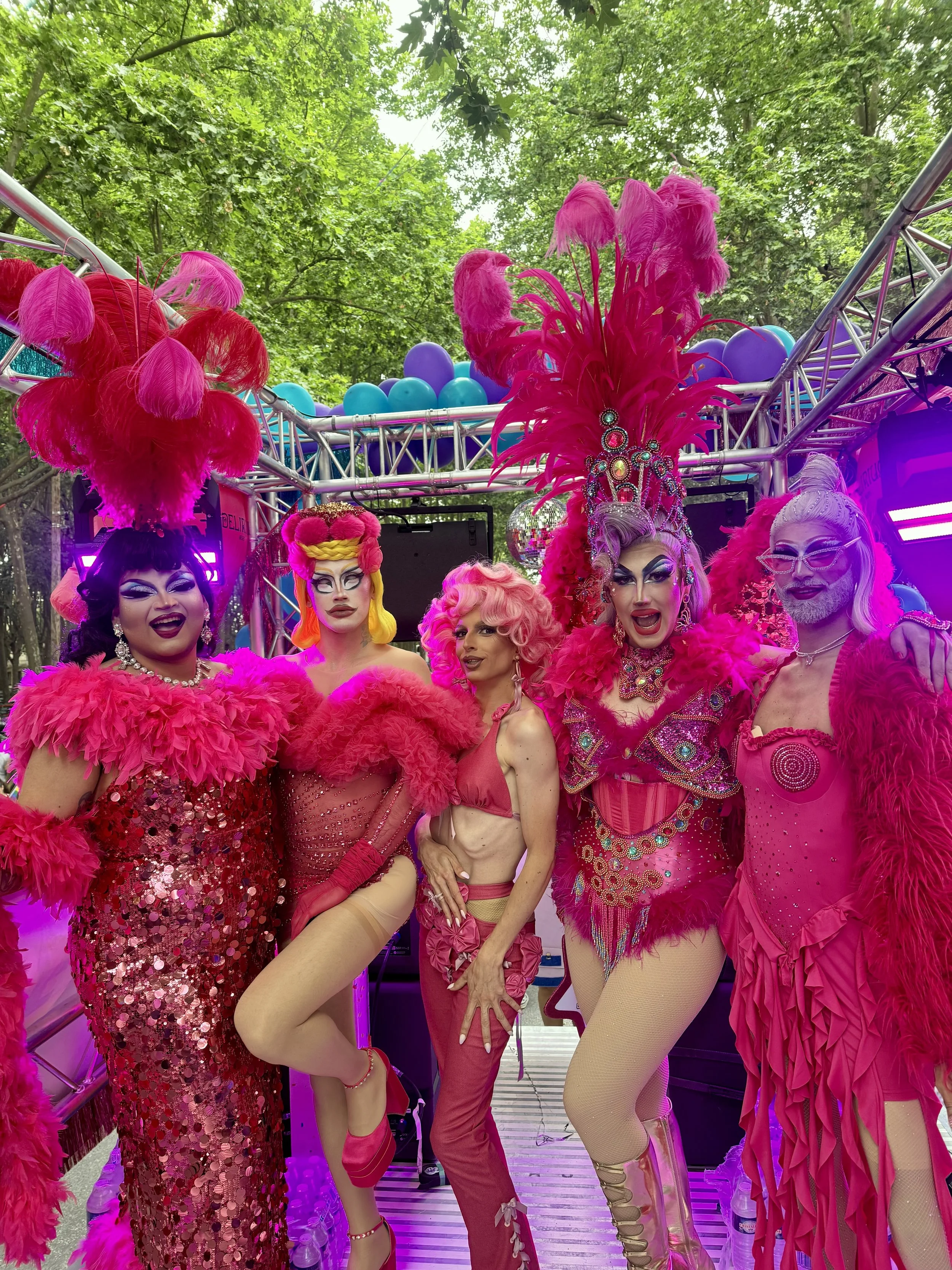 Drag queens en costumes roses, posant dans un défilé, avec des plumes, des perruques colorées et des maquillage élaboré, devant un décor en plein air avec des arbres.