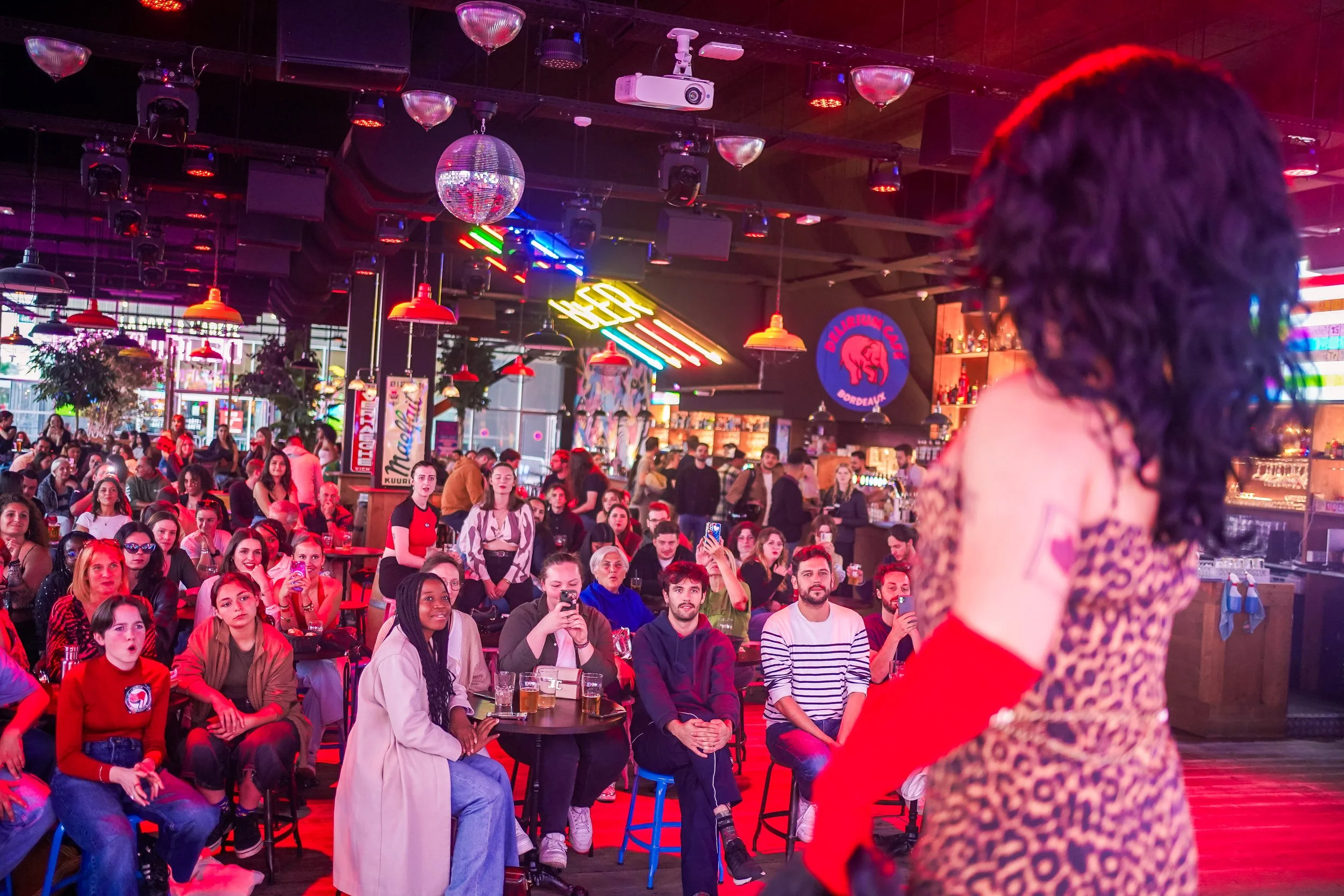Une femme qui chante ou s'adresse à un public dans un bar ou une salle de spectacle, avec une foule de spectateurs attentifs et certains prenant des photos. L'ambiance est lumineuse avec des lumières colorées et des décorations néon.