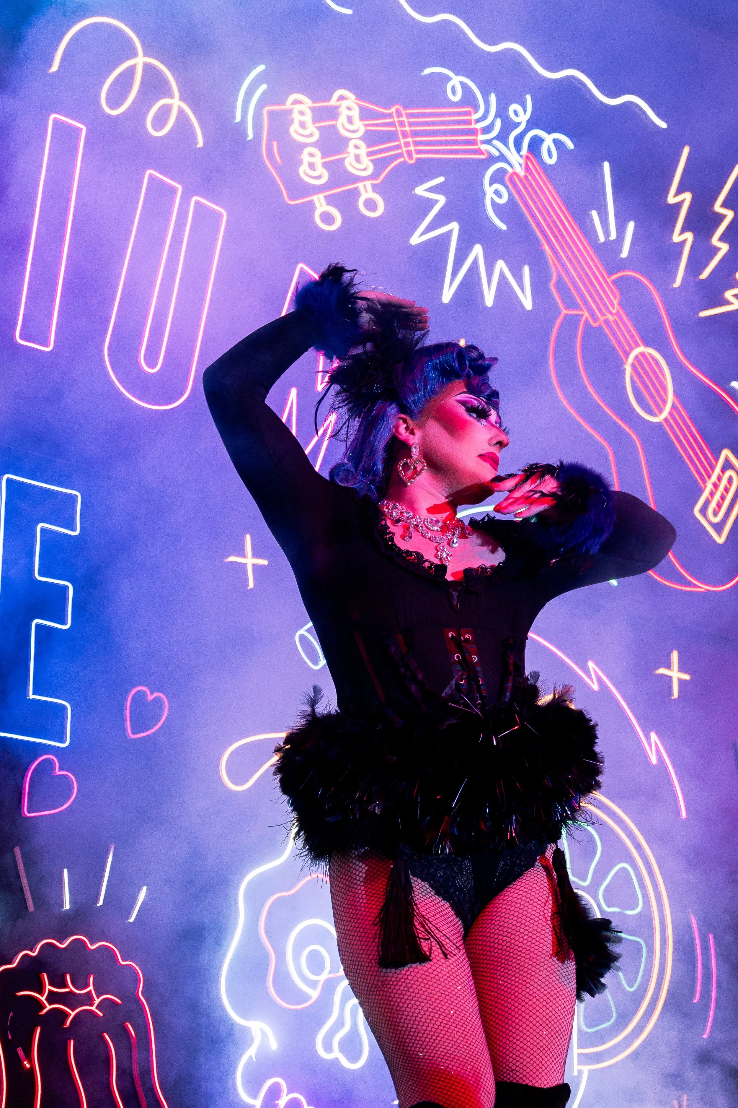 La drag queen, Kitsch, danse devant un fond avec des néons colorés représentant des icônes et des symboles variés, dans une ambiance lumineuse et artistique.