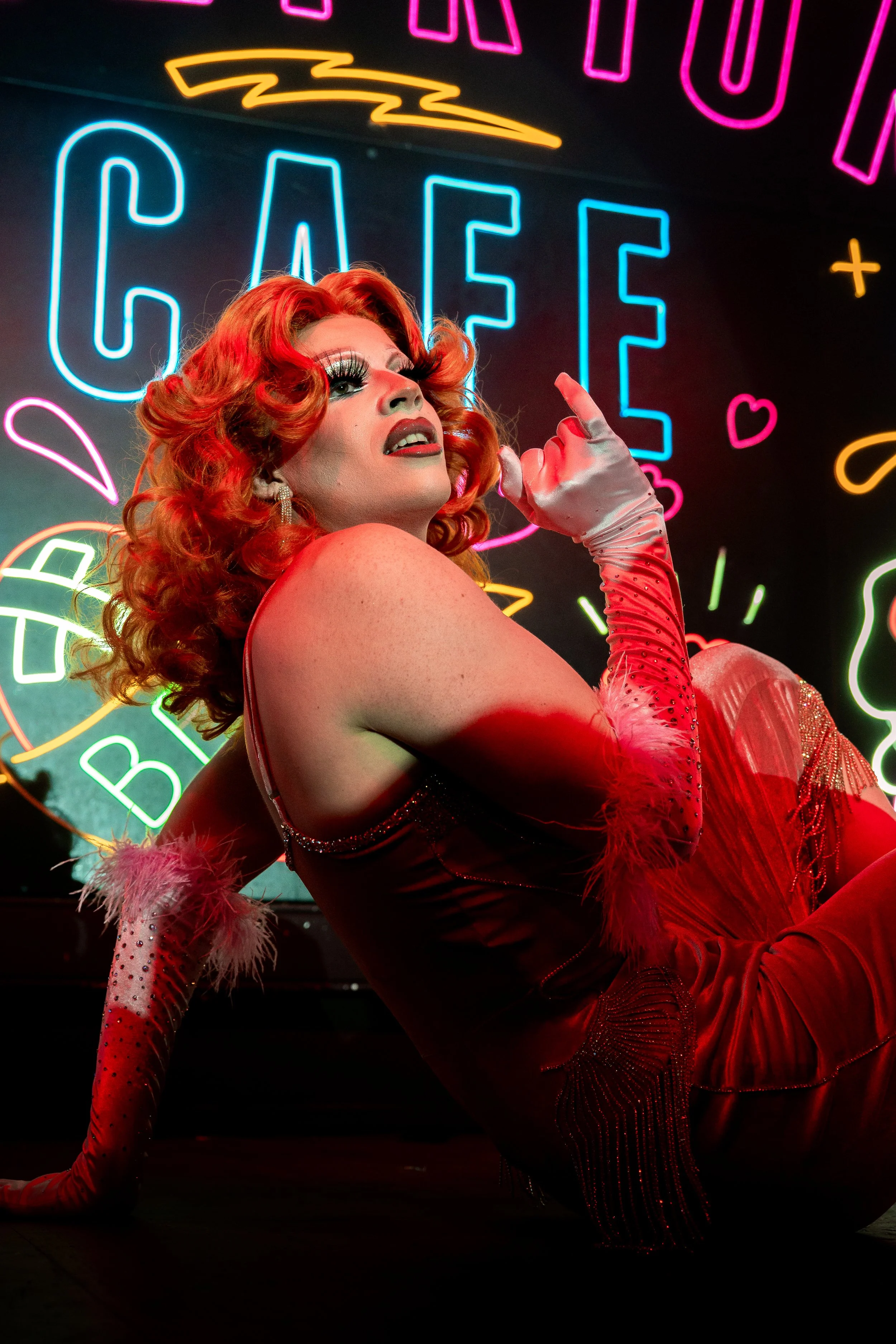 Une personne en costume de scène rouge avec des cheveux roux bouclés pose devant un panneau de néons colorés affichant le mot "CAFÉ".