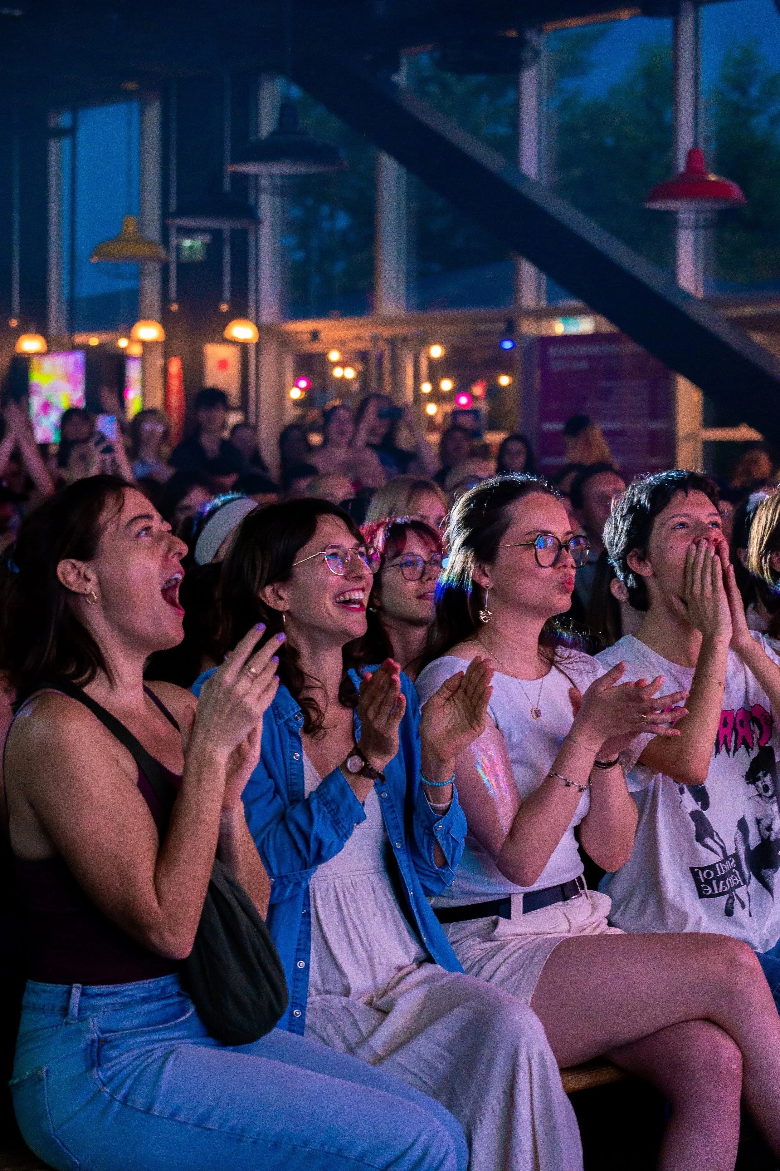 Un groupe de personnes assises, regardant et réagissant avec excitation lors du spectacle du Club Drag, dans une ambiance tamisée avec des lumières de couleur et des écrans lumineux en arrière-plan.