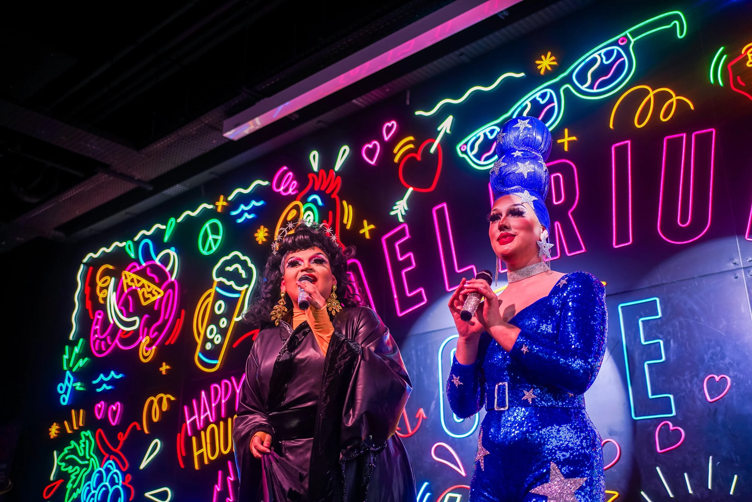 Deux drag queens chantent sous une enseigne lumineuse colorée avec des dessins de nourriture, d'icônes positives et de mots comme 'Happy' et 'Love'.