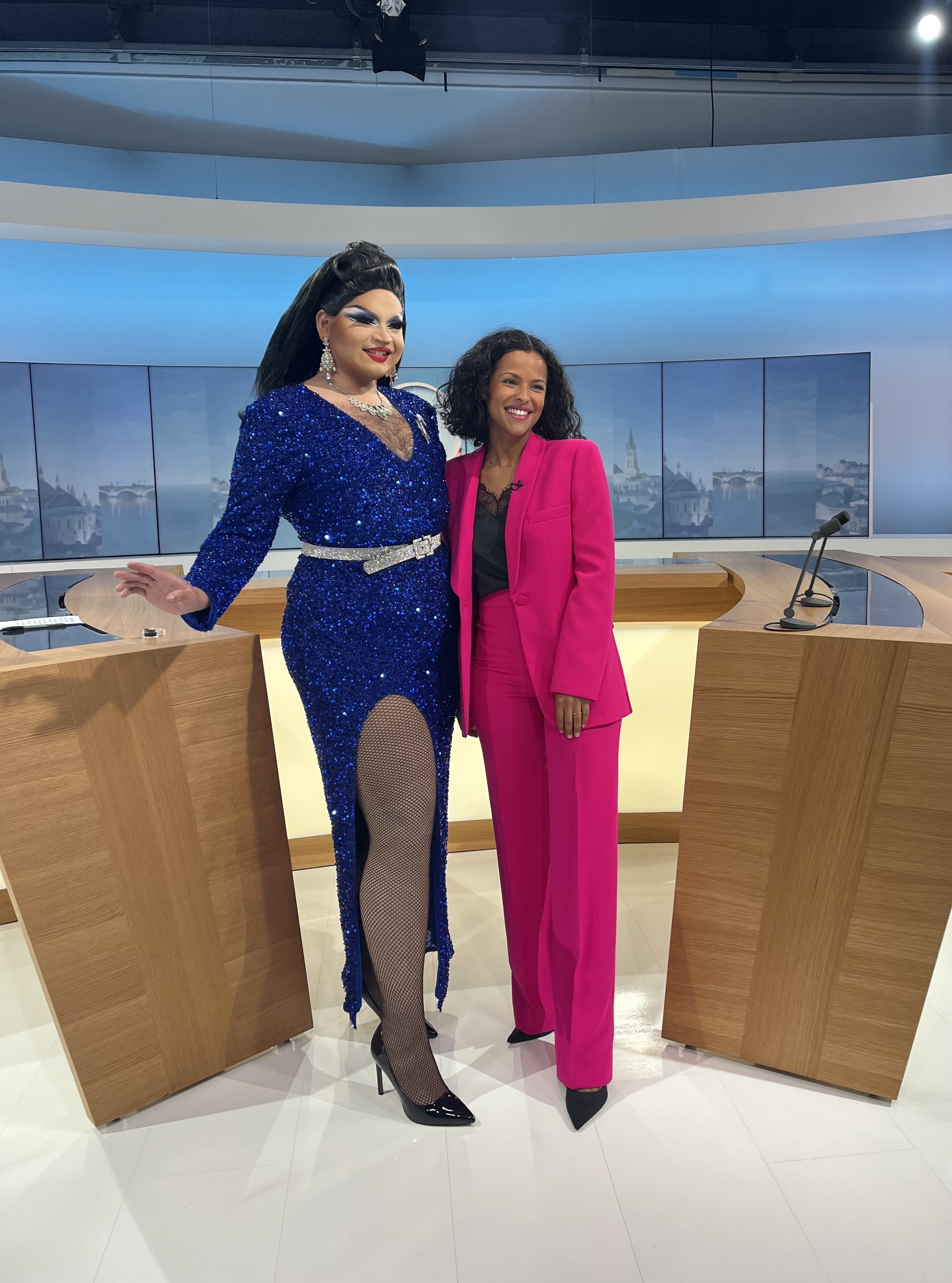 Carmen Santa Cruz, drag queen, sur le studio de télévision France 3, l'une en robe bleue brillante et l'autre en costume rose, souriantes, avec un décor de ville en arrière-plan à l'écran.