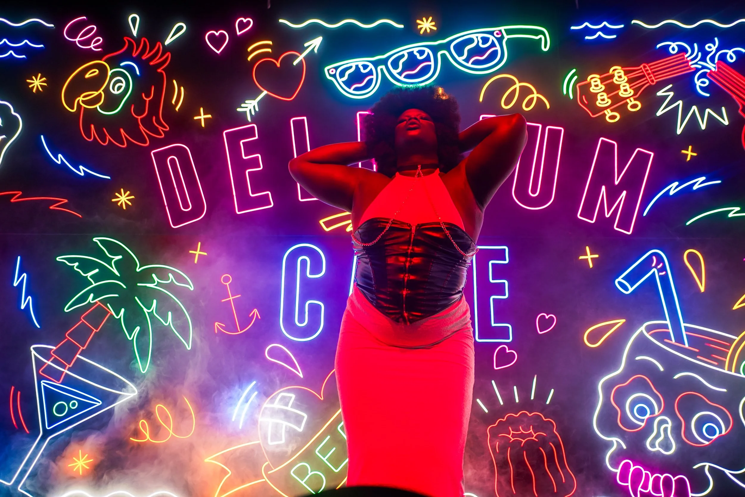 Une femme pose devant un mur illuminé par des néons colorés représentant divers motifs et mots liés à l'été, comme des visages, des palmiers, des lunettes de soleil et des cocktails.