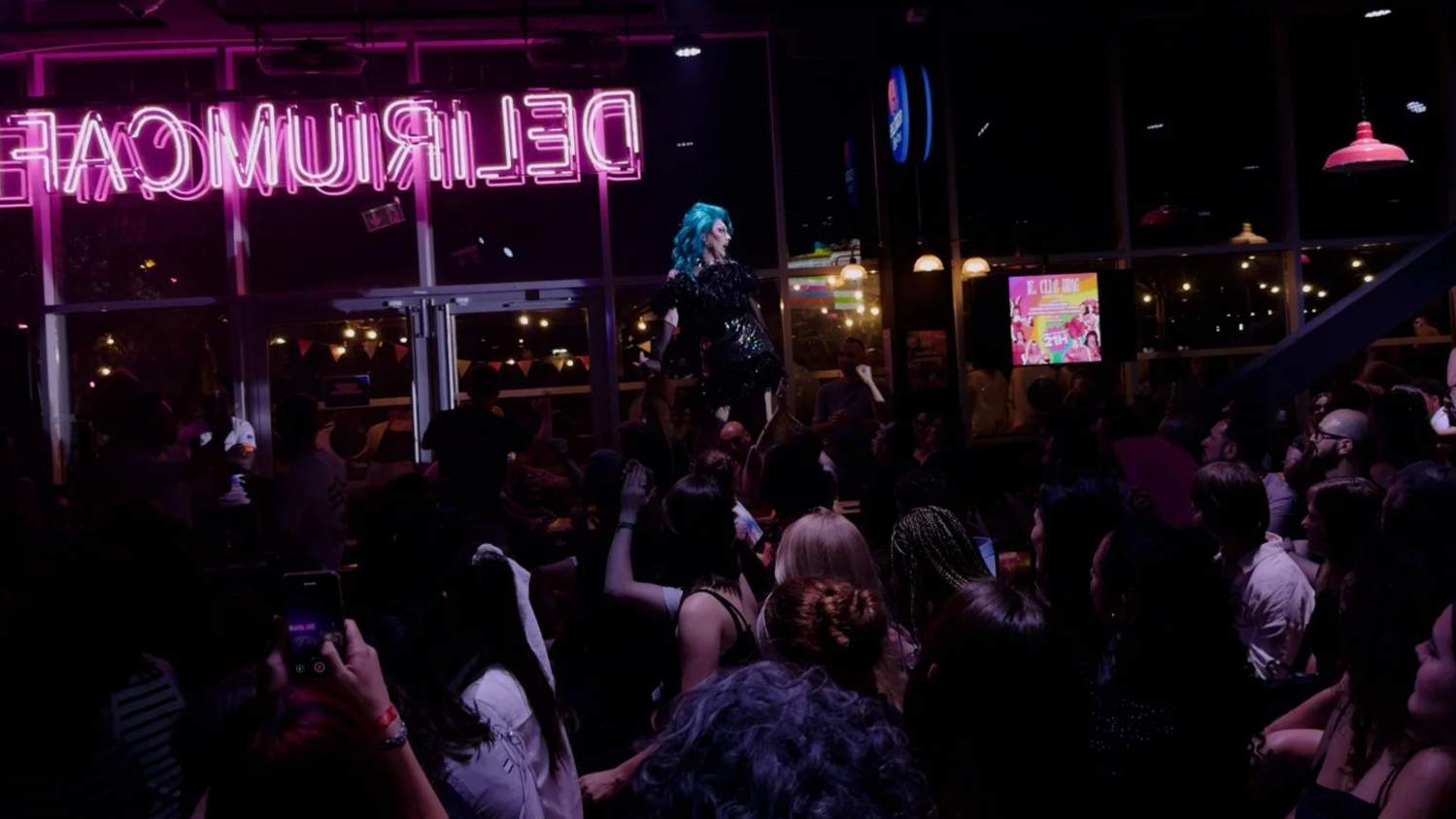 Spectacle drag avec une performeuse aux cheveux bleus et costume brillant, devant un public dans un bar avec enseigne néon rose lumineux 'DELRMIQUE'.