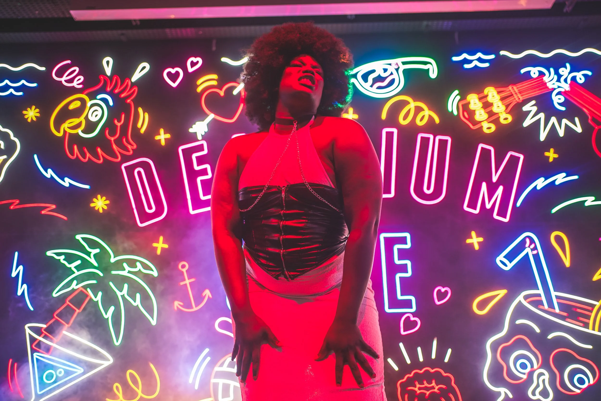Une femme avec des cheveux afro portant une robe rouge, dans un arrière-plan de néons colorés en forme de dessins de détails comme un ananas, une guitare, un visage avec des lunettes, des cœurs, des palmiers, des éclairs, et des éclaboussures de peinture lumineuses.