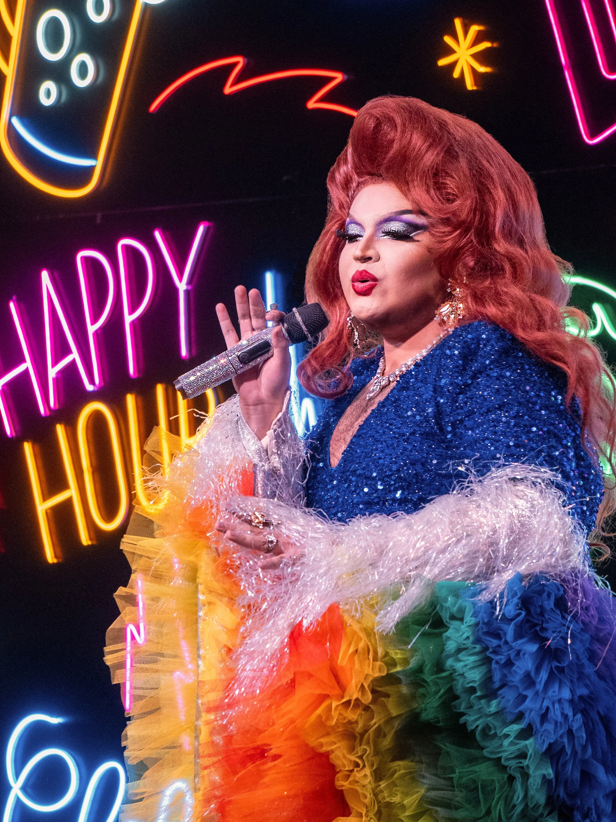 Une drag queen, Carmen Santa Cruz, porte une robe colorée en tulle et des bijoux brillants. Elle chante dans un micro devant un panneau de néons colorés avec les mots 'HAPPY' et autres motifs.