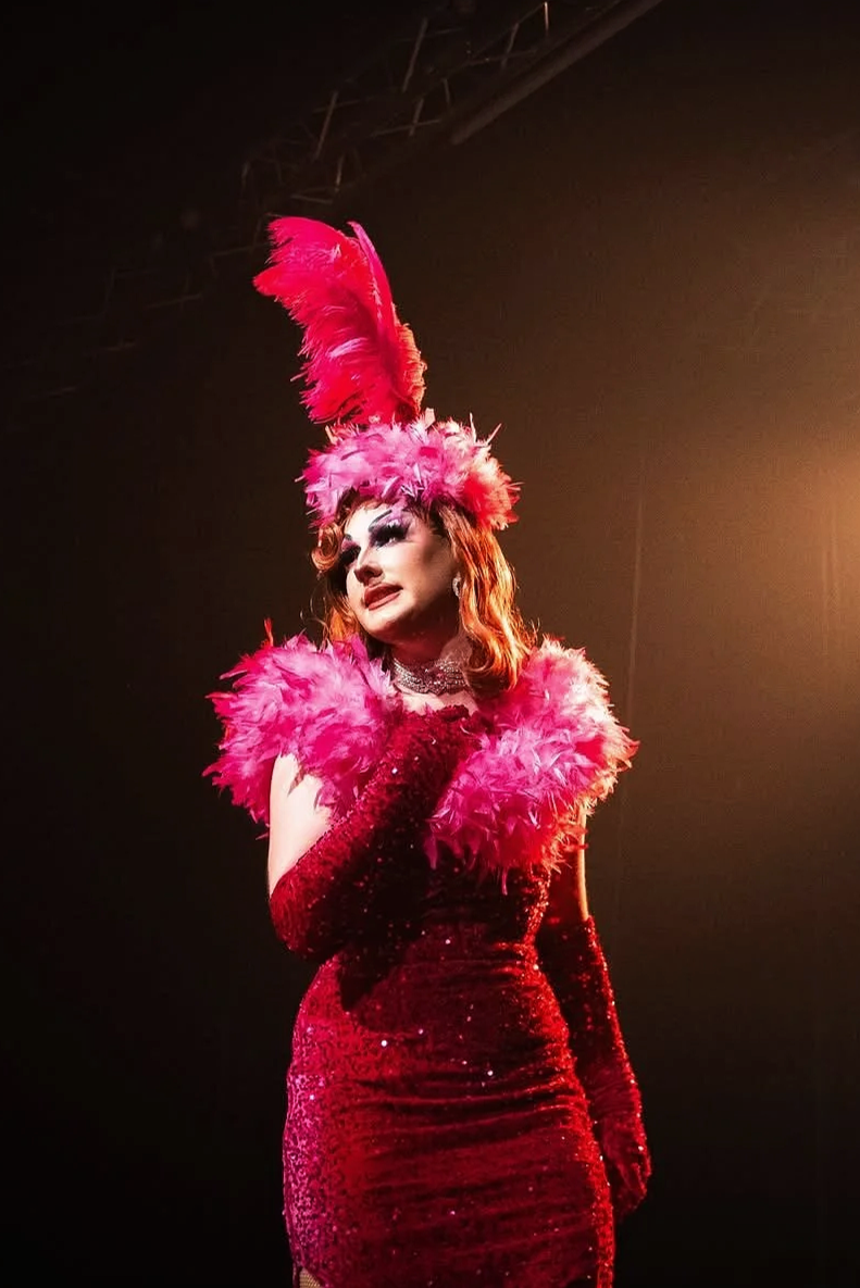 Kitsch, Drag Queen, en costume de spectacle rouge avec des plumes roses, portant une robe pailletée rouge et des gants longs, doit probablement faire une performance ou un spectacle sur scène.