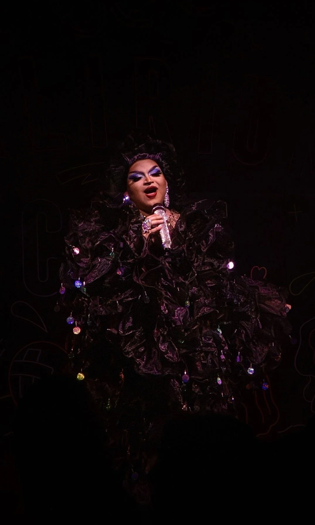 Une drag queen, Carmen Santa Cruz, chante sur scène, portant une robe noire ornée de lumières colorées, avec un maquillage spectaculaire et des boucles d'oreilles brillantes.