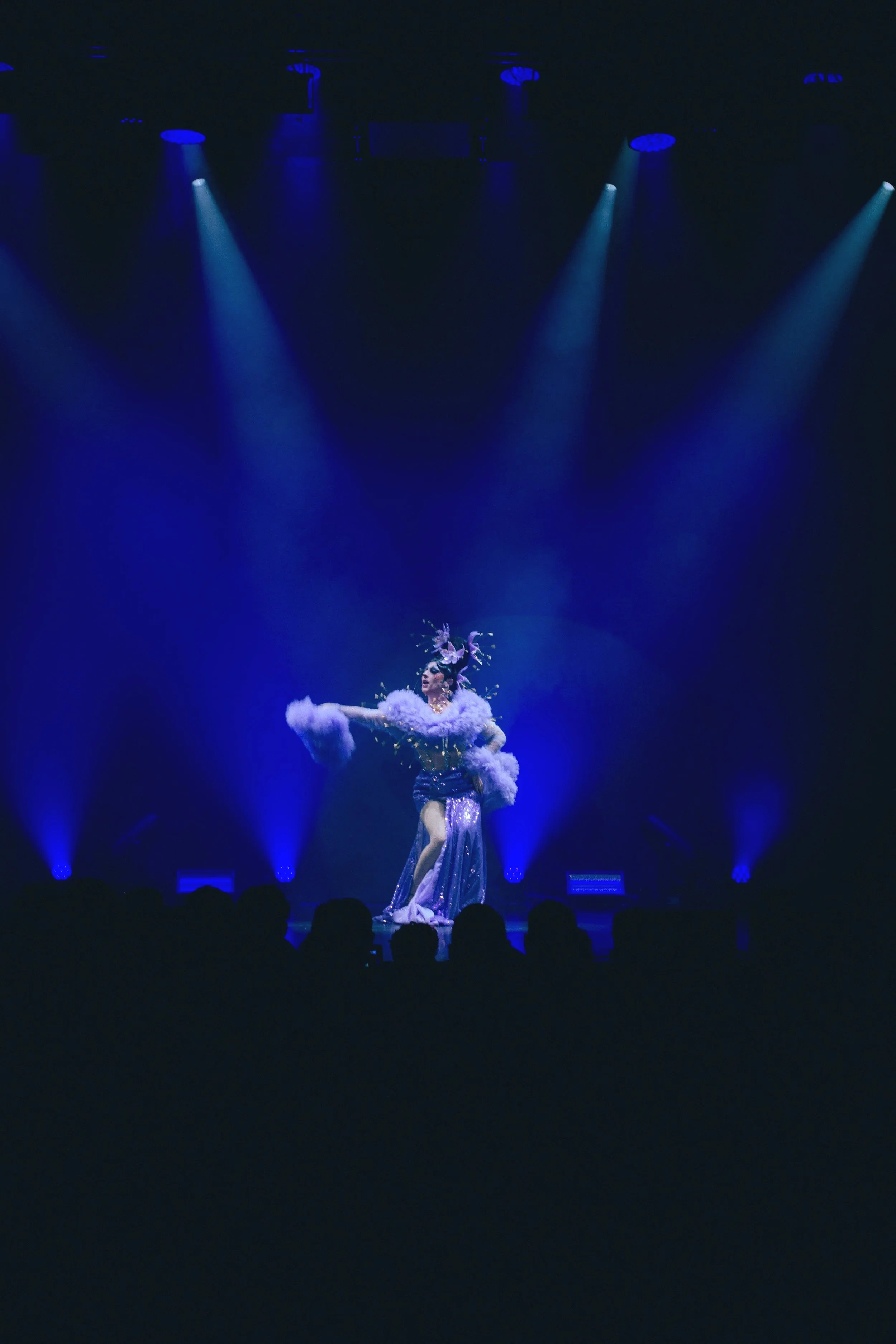 Une scène de spectacle avec une artiste en costume brillant et accessoires floraux, sous des projecteurs bleus dans une salle sombre.
